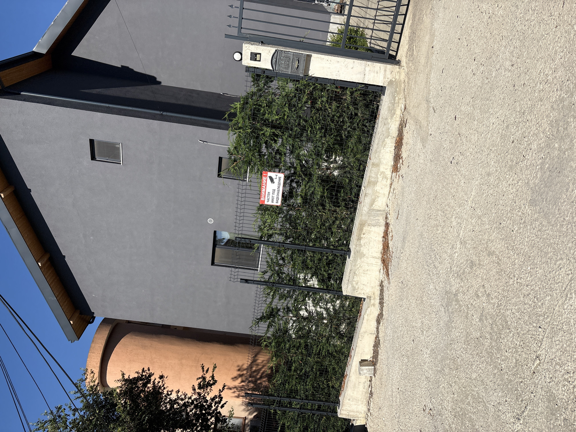 The image depicts the exterior of a modern grey house with a wooden-trimmed roof, situated in Varna, Bulgaria. Adjacent to the grey house is a tall, cylindrical, terracotta-colored structure. A black metal fence with an attached hedge runs along the front of the property. On the fence, a red and white sign reads "ВНИМАНИЕ! ЧАСТЕН ИМОТ ПОД ВИДЕОНАБЛЮДЕНИЕ" (Warning! Private Property Under Video Surveillance). A black metal gate with decorative elements stands open on the right side of the frame, next to a concrete pillar with a mailbox. The ground in front of the property is paved with concrete and appears to be a driveway or sidewalk. Power lines are visible in the upper left corner of the image, against a clear blue sky.