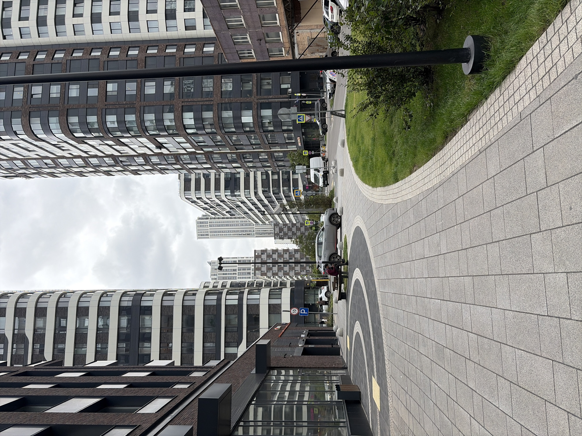 The image shows a street scene in Moscow, Russia, featuring a modern residential complex with tall, multi-story buildings. The architecture is characterized by curved facades, a mix of dark and light exterior panels, and numerous windows. A paved walkway with a patterned design curves through the foreground, bordered by a grassy area and a dark lamppost. In the background, several cars are parked along the street, including a white van and a silver SUV. A few pedestrians are visible, including one person sitting on a bench and another walking. The sky is overcast. The scene depicts an urban environment with contemporary residential architecture.