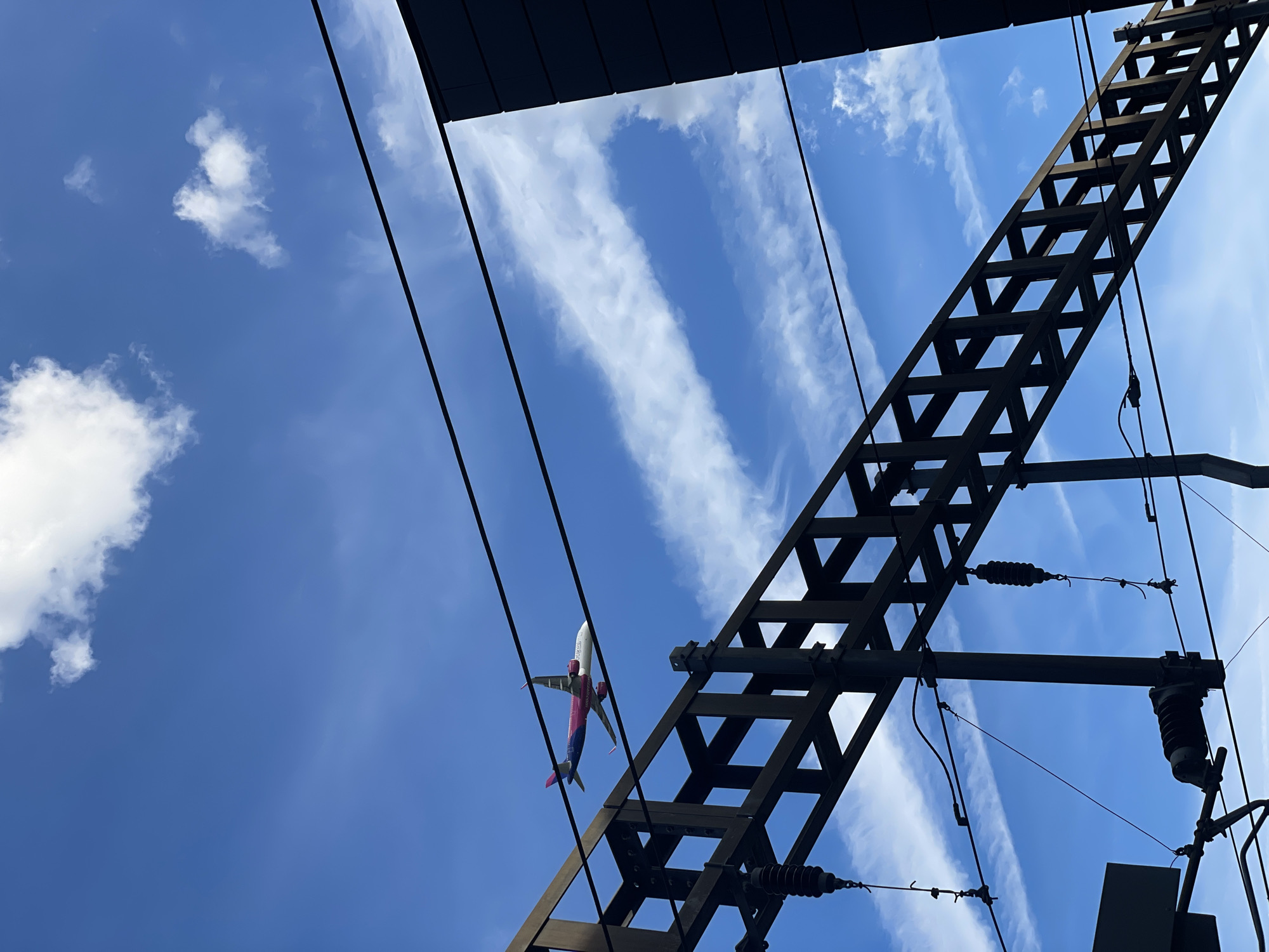 An airplane with pink and blue markings flies across a bright blue sky, partially obscured by thin, wispy clouds. In the foreground, the dark silhouette of electrical infrastructure, likely related to a railway line, dominates the lower portion of the frame. This structure consists of dark metal beams and wires, with some insulators visible. The scene suggests an outdoor, possibly urban, environment, with the sky indicating daytime. This specific scene is located near Luton Airport in Luton, United Kingdom, where air traffic is common.