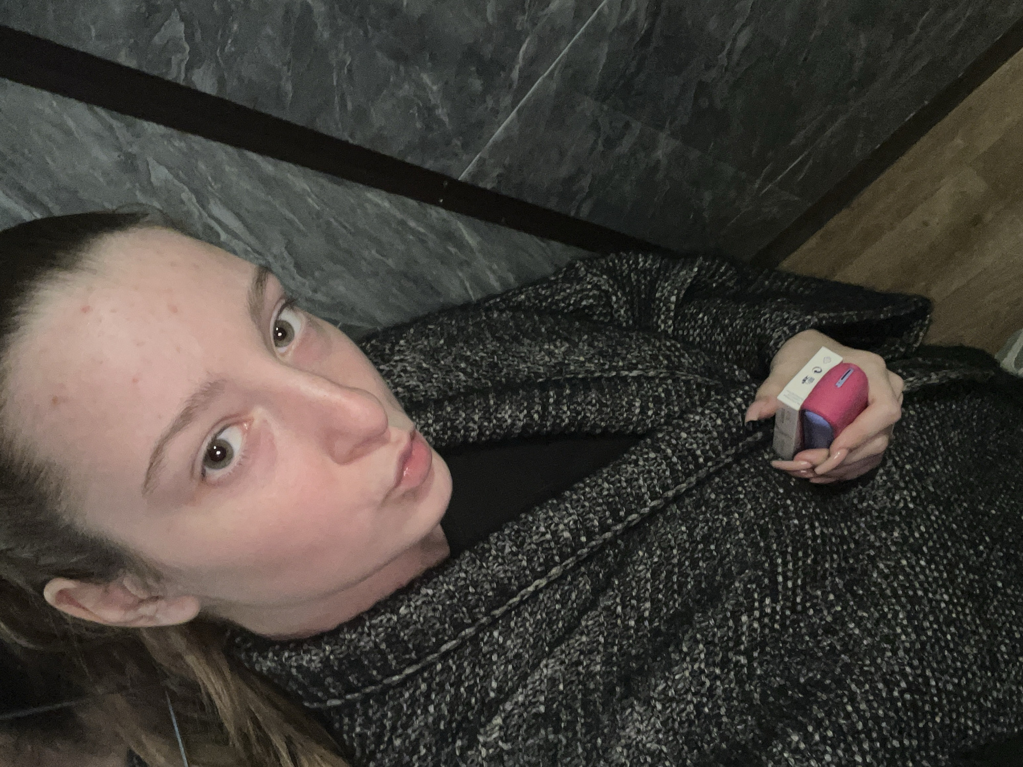 The image features a young woman captured in a medium-close-up shot, possibly a selfie, with the camera angled above her while she appears to be leaning back or lying down. She has light skin, light brown hair pulled back, and her light-colored eyes (likely hazel) are wide open, looking directly at the viewer. Her expression is a slight pout with lightly parted lips. There are a few small blemishes visible on her forehead.
She is dressed in a dark, textured, possibly knitted cardigan or jacket, worn over a darker top. In her right hand, she holds a small electronic device, which appears to be a case or charger for earbuds or a vaping device. It is two-toned, with one side being bright pink or magenta and the other white or silver. On the white section, several small regulatory symbols are visible, including what resembles a "CE" mark and a crossed-out wheeled bin icon, along with other faint, illegible text. Her fingernails are long and appear to have dark polish.
The background primarily consists of dark grey or black textured surfaces, likely large tiles or panels, positioned diagonally behind her. A thin, dark line runs across this dark background. In the lower right corner, a section of light brown wooden flooring or paneling is visible, also at an angle. The lighting indicates an indoor setting. There are no specific cues within the image to determine the time of day or weather. While the location is contextualized as Varna, Bulgaria, the image itself does not contain specific geographical identifiers for that city. The overall scene is a casual, direct portrait.