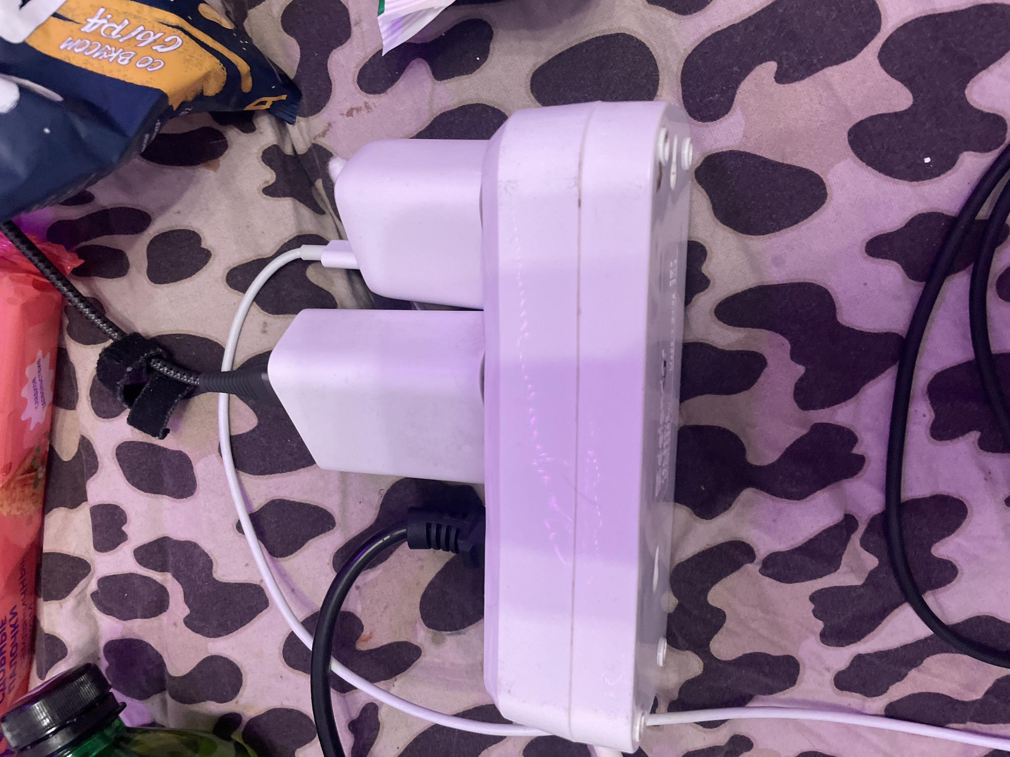 A white power strip is plugged into an electrical outlet and is covered with a cow-print fabric. Three white adapters are plugged into the power strip. Two black power cords and one white power cord are plugged into the adapters. A green bottle is to the left of the power strip. A bag of chips is to the right of the power strip. The image is taken from a top-down perspective. This scene is not located in Irinovka, Russia.