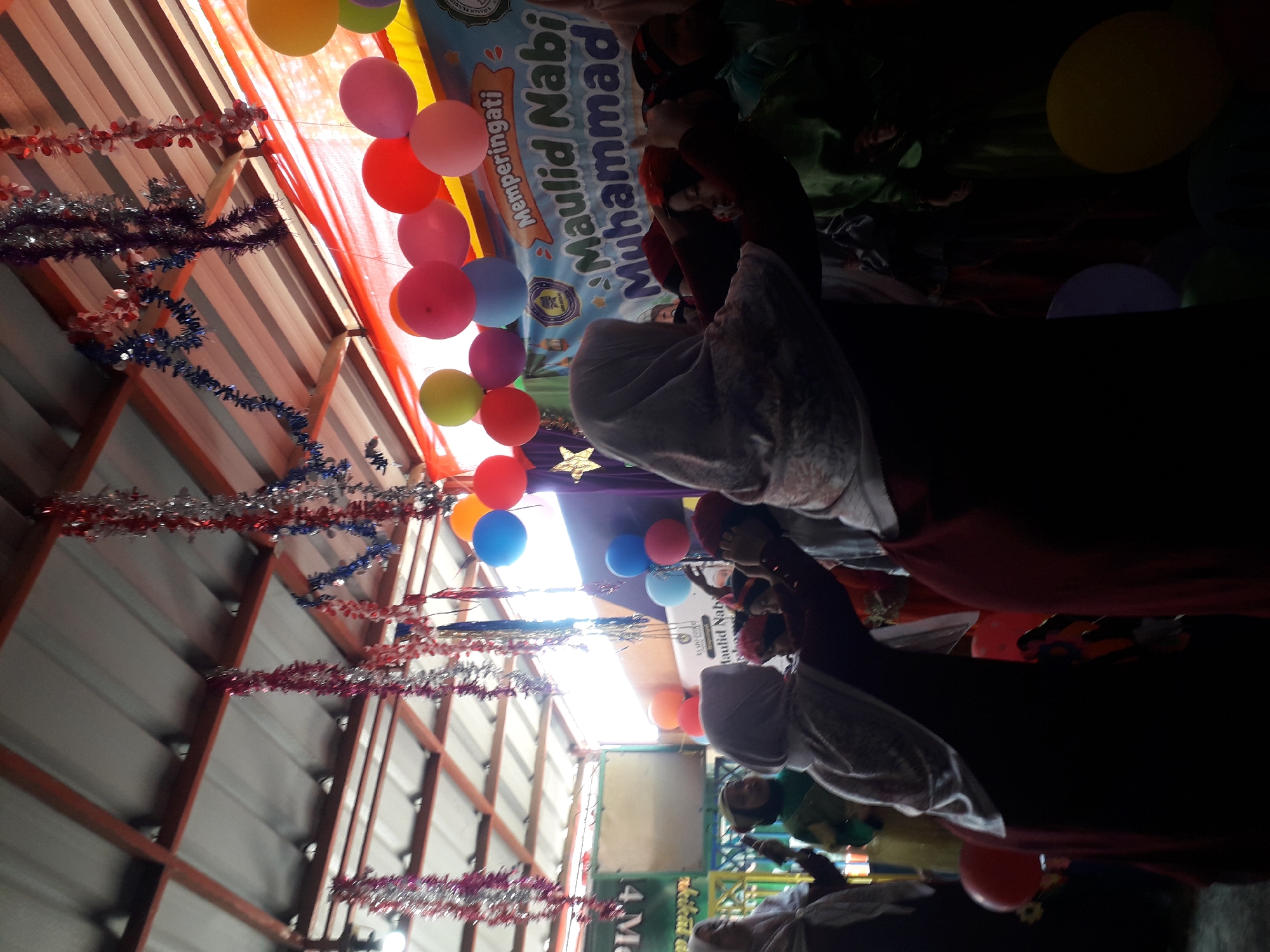 In the city of Cankuang, Indonesia, a festive gathering is underway, likely celebrating Maulid Nabi Muhammad. A group of individuals, predominantly women and children, are present under a makeshift outdoor roof structure adorned with red, white, and blue tinsel garlands. Balloons in various colors, including yellow, orange, pink, and blue, are scattered throughout the scene, some attached to a banner that reads "Memperingati Maulid Nabi Muhammad" in Indonesian.
The foreground is dominated by the silhouettes of two women in Islamic attire, one in a dark maroon garment and the other in a dark brown garment, both wearing hijabs. The woman on the left appears to be interacting with a child, adjusting a red headdress on the child's head, who is wearing a green outfit. Another woman in the background, also in a hijab, is seen holding a cell phone. The atmosphere suggests a communal celebration, with people gathered for an event of religious significance. The lighting is dim, with the bright outdoor sunlight filtering through the canopy and illuminating some of the balloons and banners, creating a high-contrast scene.