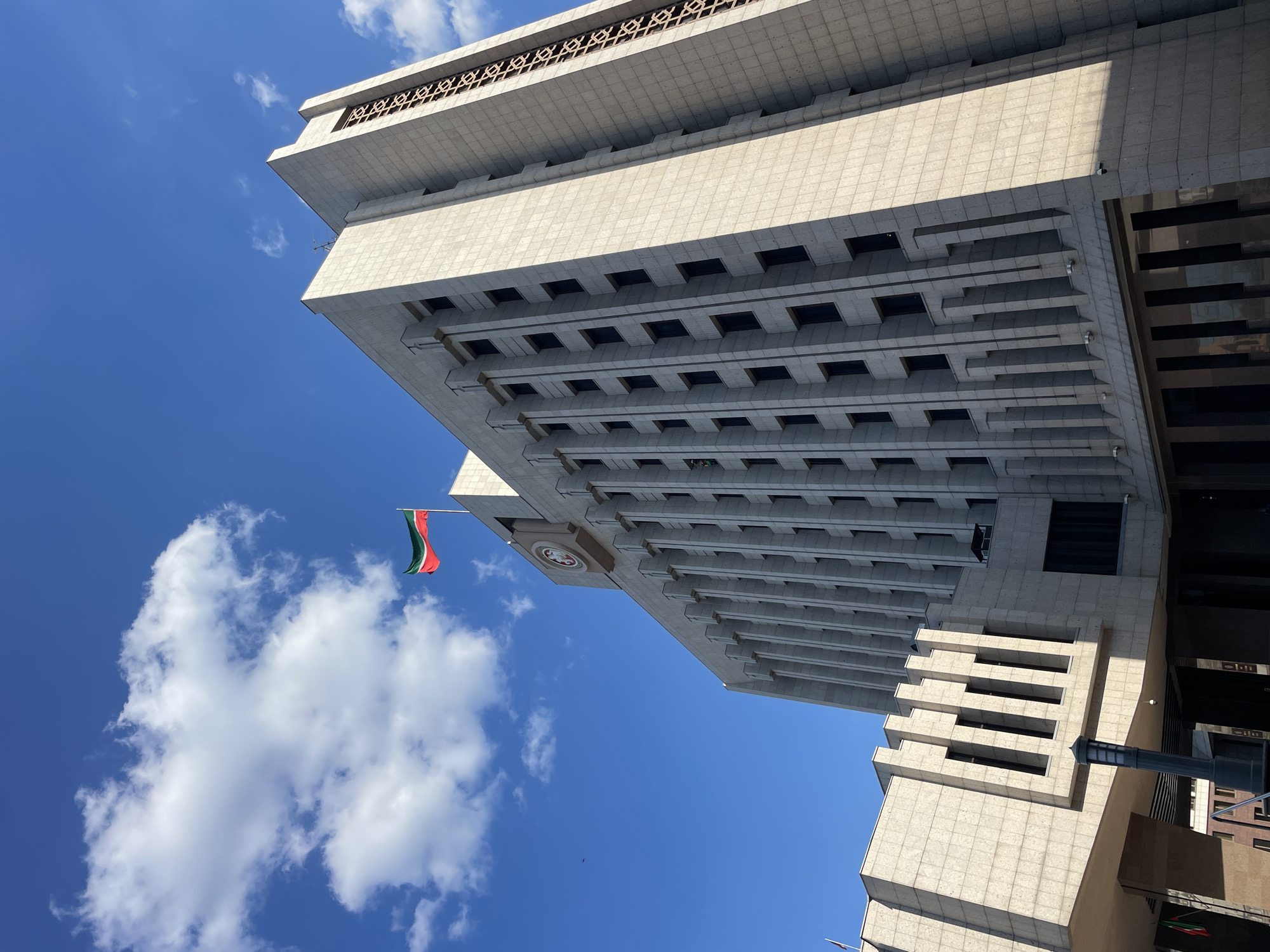 A low-angle shot depicts a multi-story building in Unknown, Unknown, against a blue sky with scattered white clouds. The building is constructed from light-colored stone or concrete and features a pattern of recessed windows across multiple floors. A flag with green, white, and red stripes hangs from a flagpole extending from the building's facade. The building's architecture includes tiered sections and a decorative, lattice-like element at the top. A street lamp is visible at the lower right of the frame.