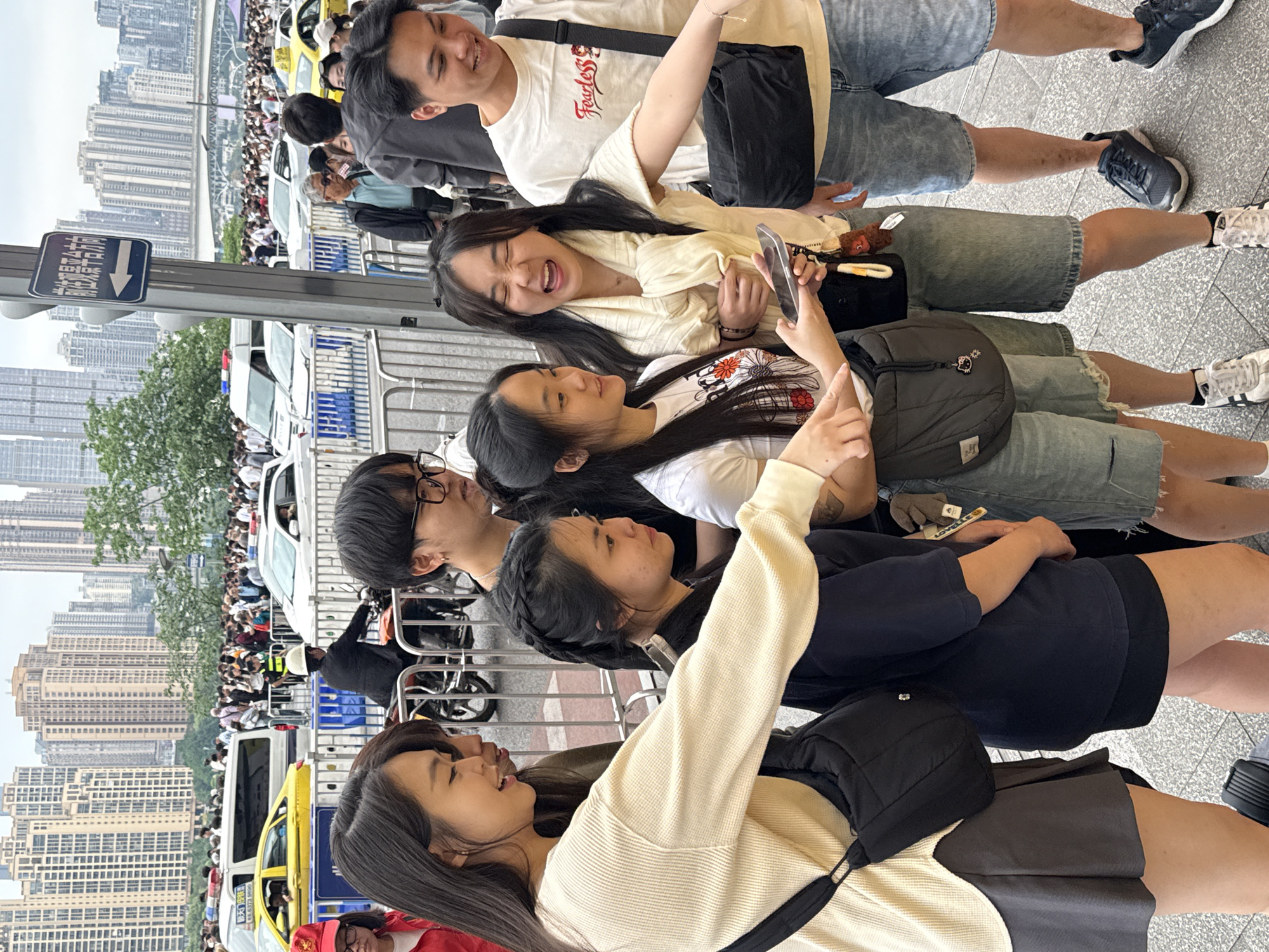 The image captures a vibrant urban scene, likely in Yuzhong District, China, during daylight hours under an overcast sky. In the foreground, a group of seven young Asian adults, predominantly women, are engaged in social activity. Most are smiling or laughing joyfully, interacting with each other and their smartphones, suggesting they are taking selfies or photos. They are casually dressed in t-shirts, shorts, and light jackets or cardigans, some wearing sneakers. Accessories include shoulder bags, fanny packs, and eyeglasses.
In the mid-ground, a large crowd of people is visible, gathered behind metal barriers alongside a street. Several vehicles, including white cars, a yellow taxi, and a white police-style van with a blue light, are interspersed within the crowd.
The background features a dense urban skyline dominated by numerous high-rise buildings, characteristic of a modern city. A blue directional sign with white text and an arrow pointing to the right is prominent on the left side of the frame. The Chinese characters on the sign read "来福士广场" (Raffles City Plaza) and "轨道交通" (Rail Transit or Metro), providing clear location cues. The overall atmosphere is one of a busy public space, possibly a popular gathering spot or a queue for an event, filled with lively social interaction.
