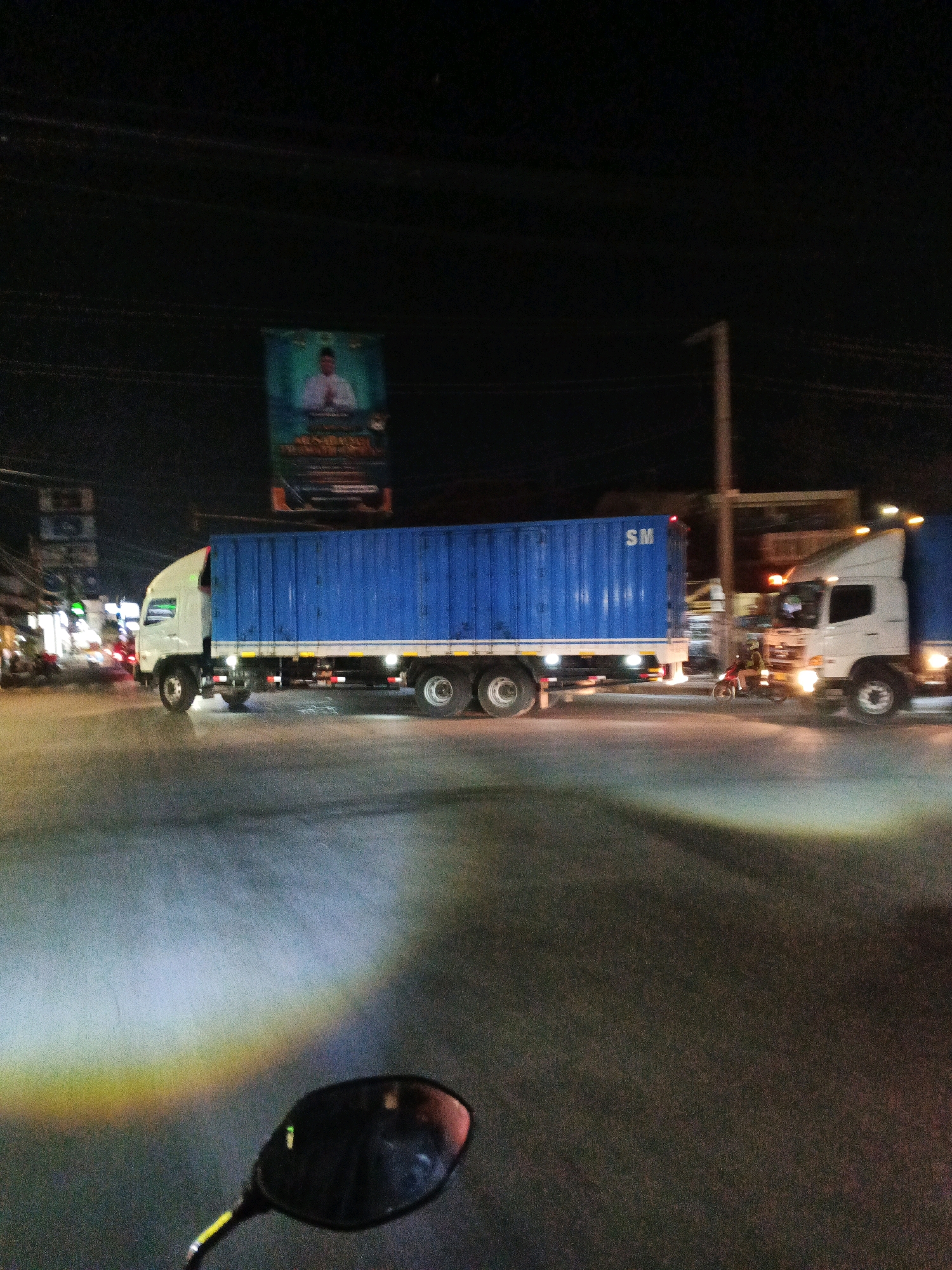 An outdoor, low-light, eye-level, horizontal shot captures a street scene at night. A white truck with a blue shipping container is the primary subject, positioned in the center of the frame. The container has "SM" printed on its side in red letters. To the right of this truck, another white truck is visible, partially obscured. A motorcycle with a rider is positioned between the two trucks, illuminated by headlights. In the background, a billboard with a man in a white shirt and a head covering is visible, along with streetlights and buildings. The foreground shows a street surface with a rainbow-like light reflection, suggesting it might be wet from rain, and a motorcycle's side mirror. The scene appears to be a busy intersection in a city.