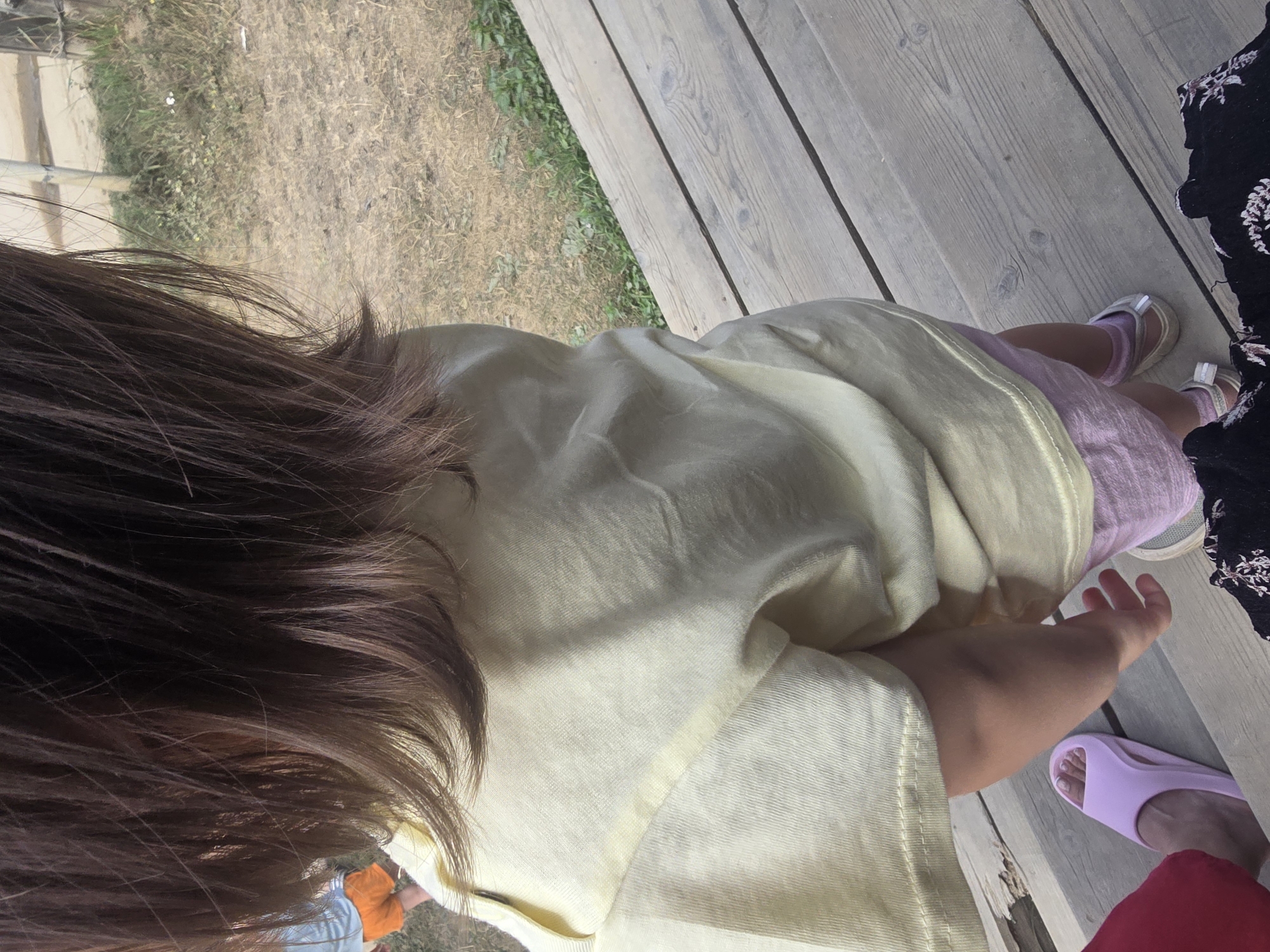 The image captures a rear view of a child with brown hair standing on a wooden deck. The child is wearing a light yellow t-shirt and pink shorts. In the background, dry grass and some green vegetation are visible. To the left, a person's foot wearing a pink slide is visible on the deck. The scene appears to be outdoors in a rural or natural setting, possibly near Maksimiha, Russia, given the presence of dry, sparse vegetation.