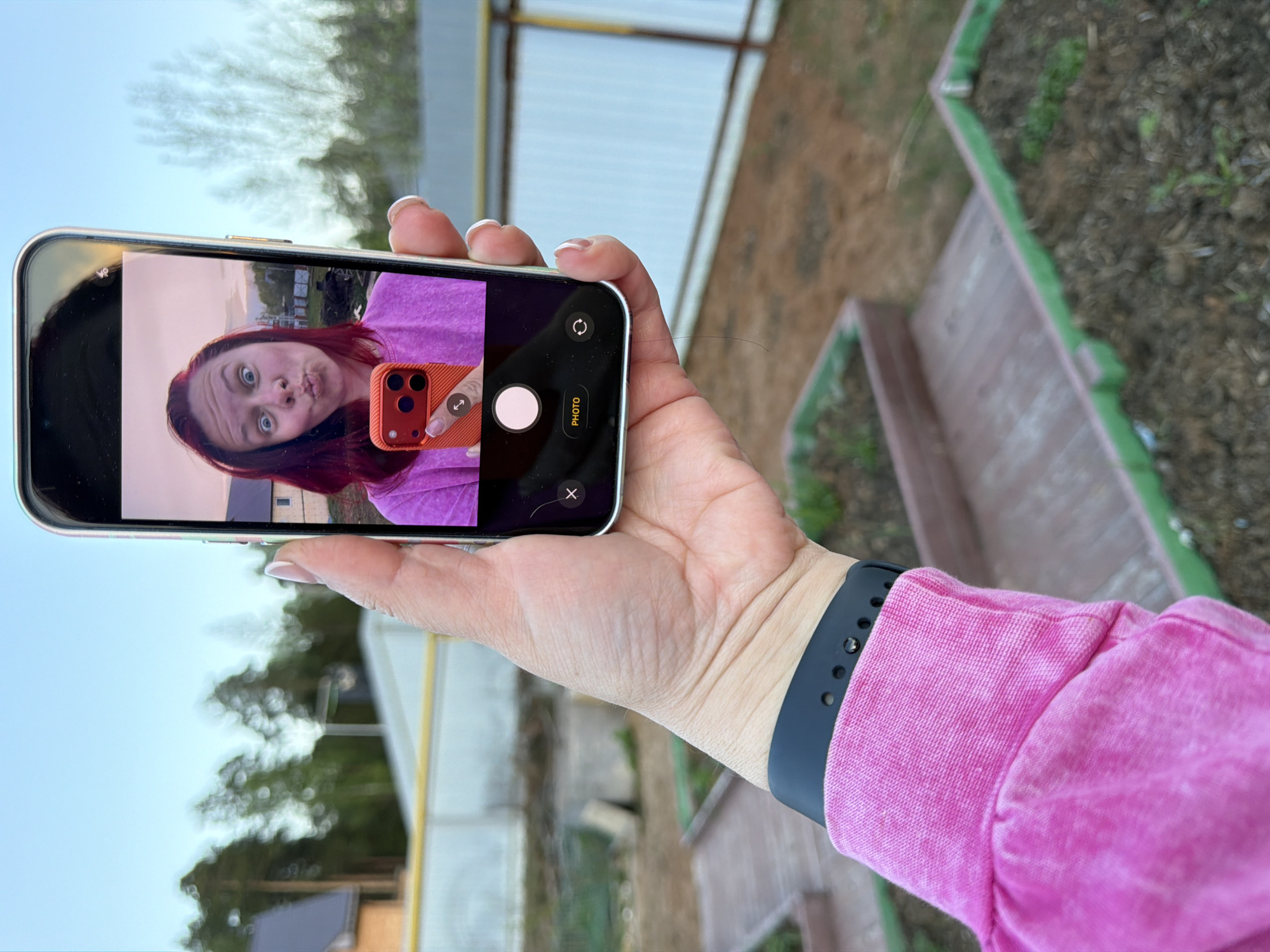 A person wearing a pink jacket and a black smartwatch holds a smartphone, displaying a selfie. The person in the selfie has red hair and is making a face. The background includes raised garden beds, a structure with white walls and yellow poles, and trees. Based on the appearance of the buildings and flora, the image may have been taken in a rural or suburban environment of Chaikovsky, Russia.