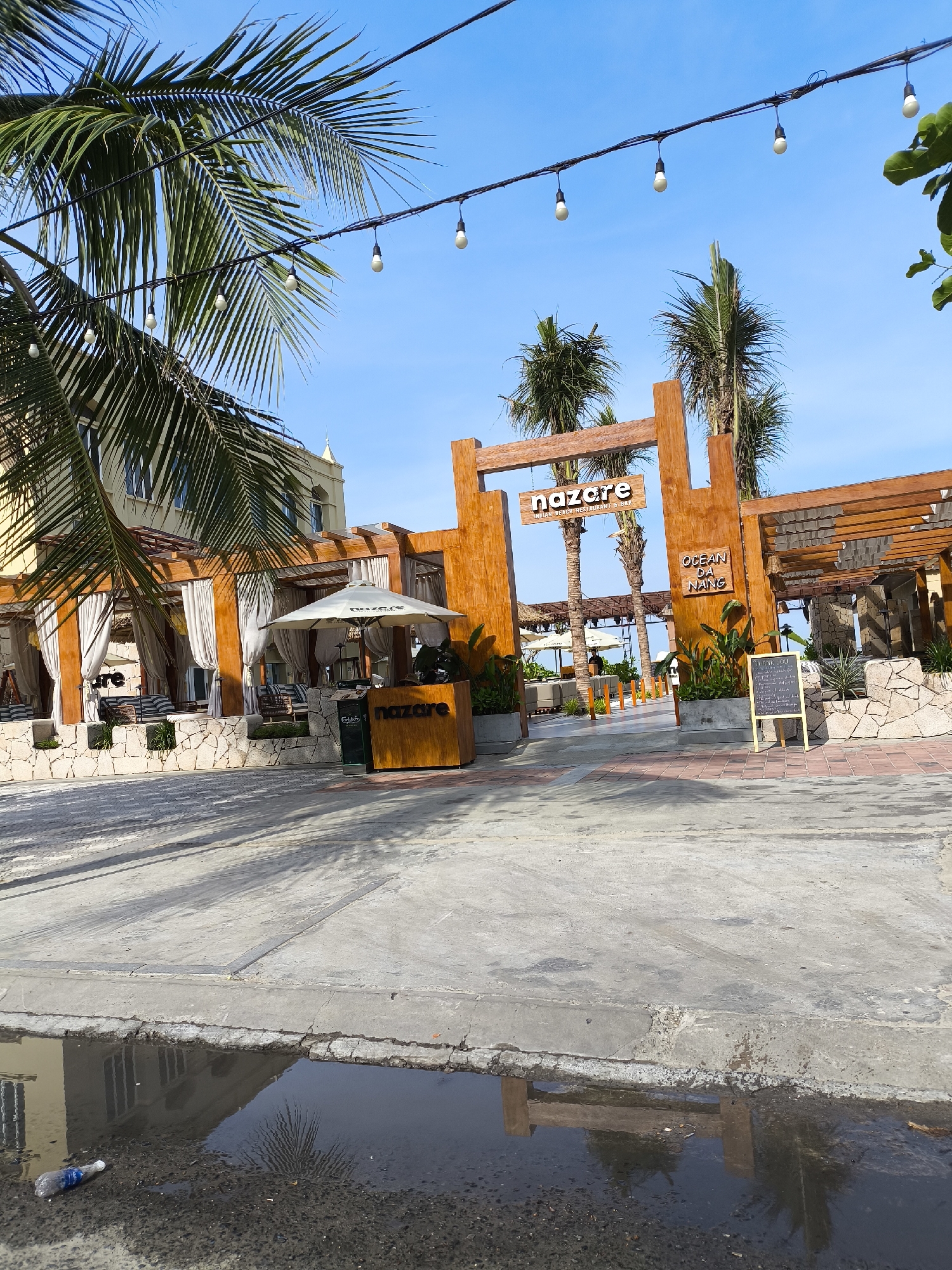 The image shows the entrance to "Nazare Indian Beach Restaurant & Bar" in Da Nang, Vietnam. The establishment features a modern, open-air design with a wooden facade, stone walls, and white drapes covering seating areas. Palm trees and overhead string lights add to the tropical ambiance. A sign identifies the location as "Nazare" and "Ocean Da Nang." The foreground shows a paved area with some puddles reflecting the sky and palm trees. A plastic water bottle is visible in one of the puddles.