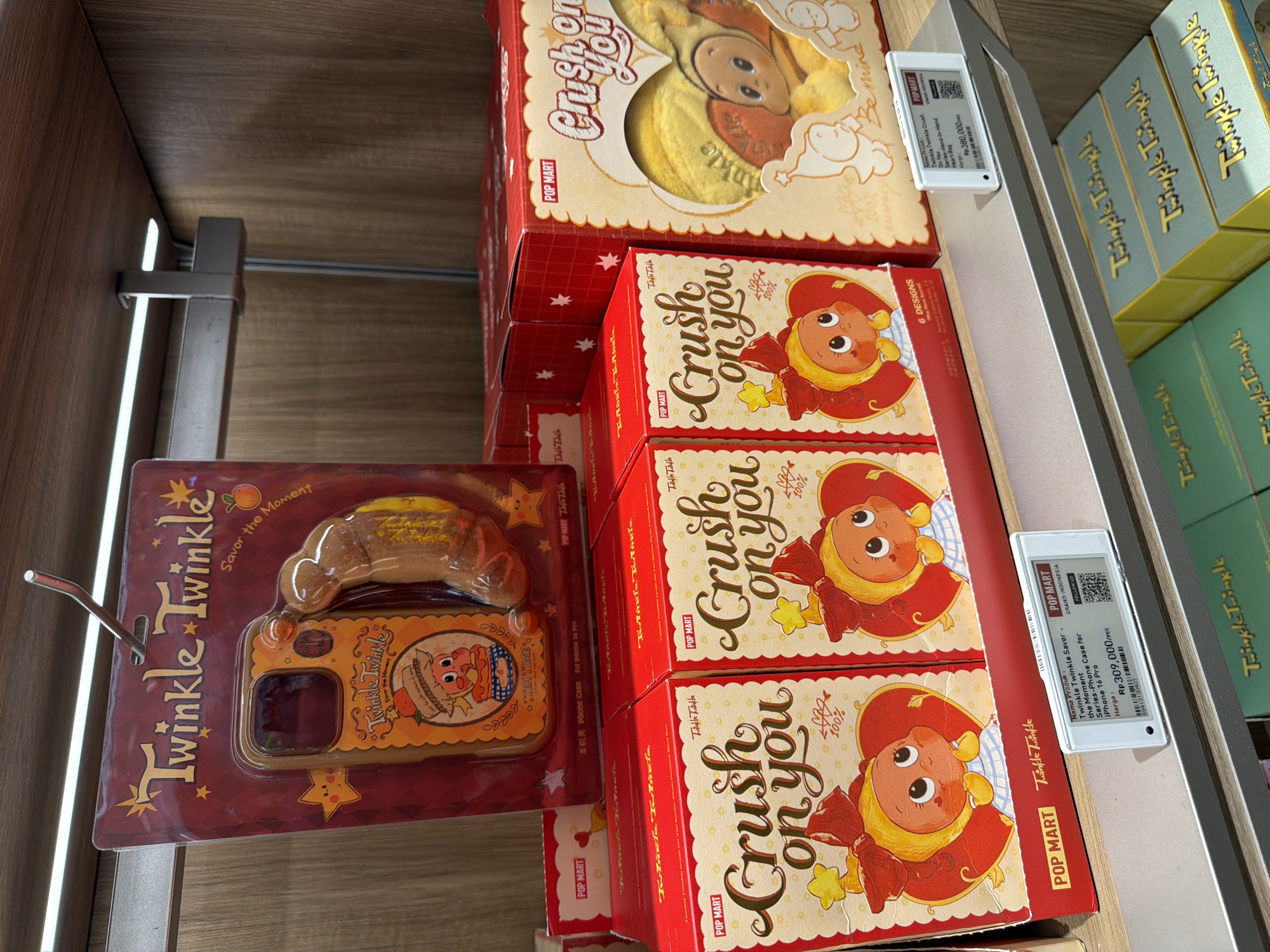 The image shows a retail display shelf at a Pop Mart store in Central Jakarta, Indonesia. The shelf is stocked with various collectible items from the "Twinkle Twinkle" and "Crush on You" series. In the foreground, several red boxes with "Crush on You" branding are visible, each featuring a cartoon illustration of a child-like character with a large red bow. A price tag indicates a price of Rp 309,000/pcs for a "Twinkle Twinkle Savor - the Moment Series-Phone Case for iPhone 13 Pro". Further back, a blister pack containing a "Twinkle Twinkle" phone case with a character and a sausage-shaped charm is displayed. To the right, a larger, heart-shaped box displays a plush toy with the "Crush on You" branding. The overall environment is that of a modern retail store, with clean shelves and lighting.