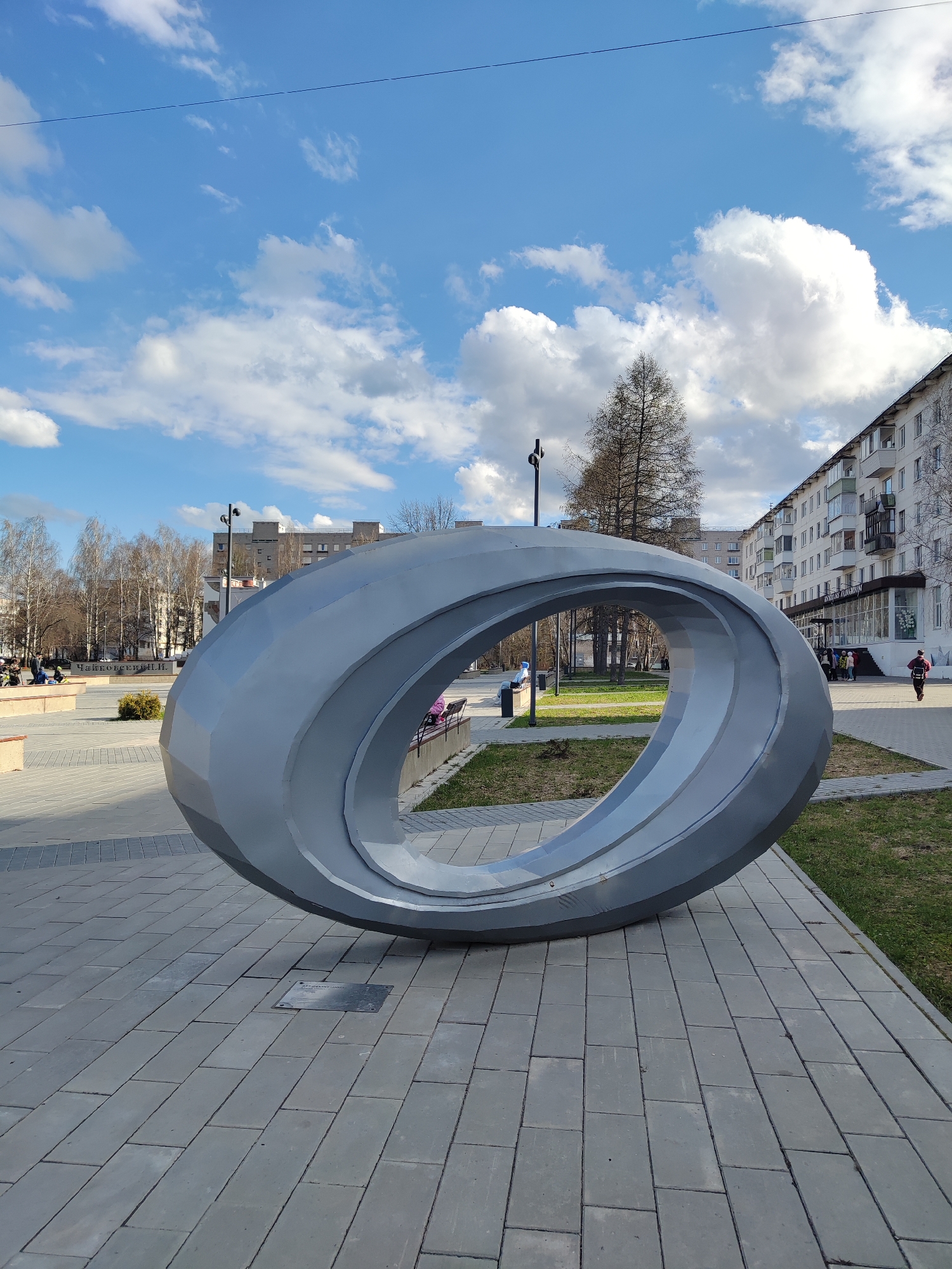 A large, modern, grey sculptural ring dominates the foreground of a public square in Chaikovsky, Russia, on a bright, partly cloudy day. The sculpture is a faceted, Mobius-strip-like form, made of metallic or painted panels, resting on a grey paved surface of rectangular blocks. A small, dark plaque is embedded in the paving near its base, bearing the Russian text "АВТОРСКИЙ НАДЗОР" (Author's Supervision).
The square features grassy patches and various types of trees, some deciduous and bare, others beginning to show early spring foliage. In the middle ground to the left, people are sitting on benches. Further back, another plaque or sign on a low concrete barrier clearly displays the Russian text "ЧАЙКОВСКИЙ". Lampposts are scattered throughout the square.
On the right side of the image, a multi-story residential building with balconies stretches into the background. A group of children and an adult are visible near its entrance, while another person walks away from the building. The sky is bright blue with scattered white cumulus clouds, and a thin overhead wire is visible across the upper portion of the frame. The overall scene depicts a calm, ordinary day in a city park or public space.