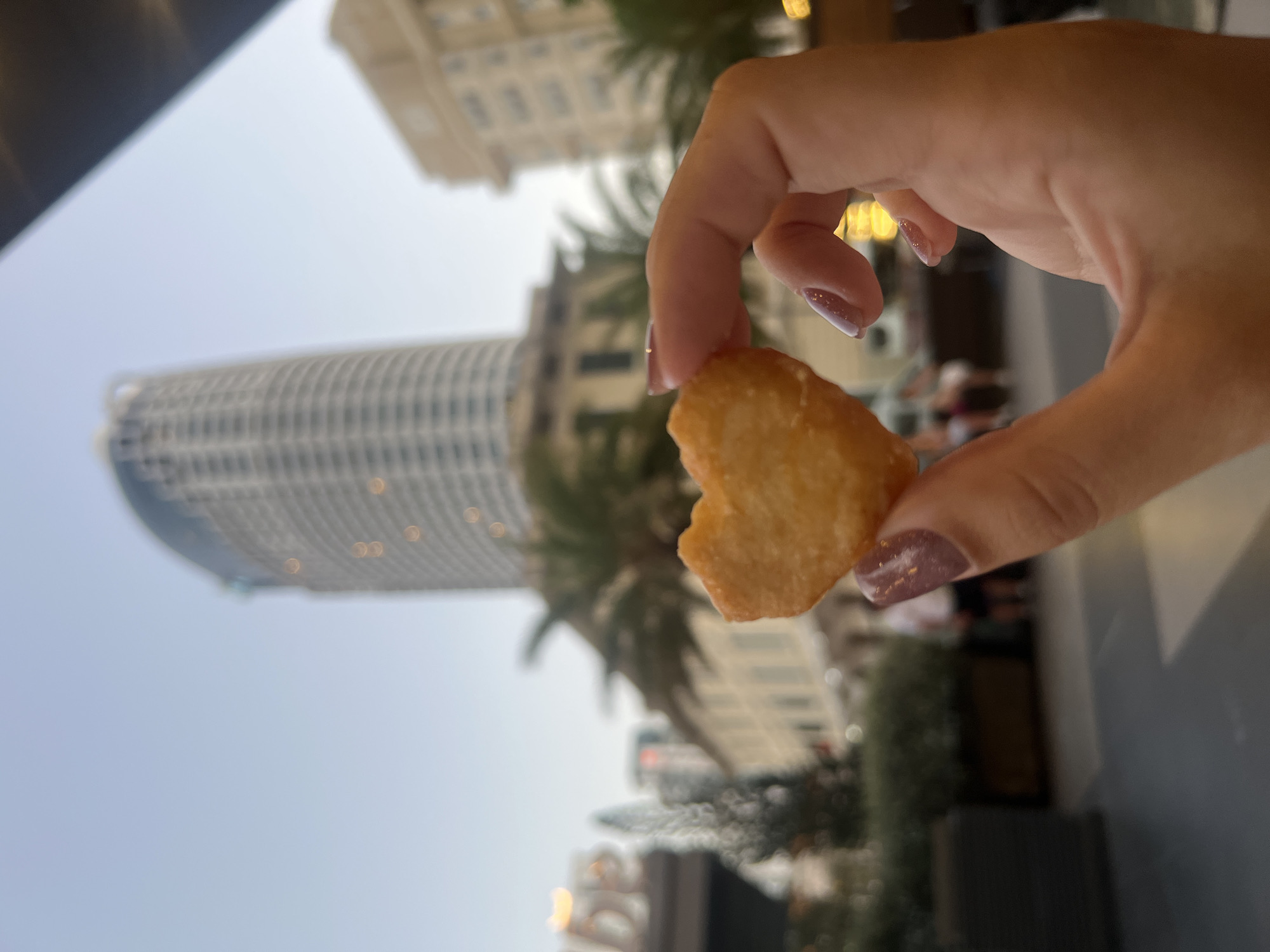 A human hand with light pink glitter nail polish on the fingernails holds a single heart-shaped, golden-brown fried food item between the thumb and forefinger in the foreground. The background is significantly out of focus, depicting an urban outdoor scene during late afternoon or early evening.
A prominent, tall, multi-story building with a curved, cylindrical facade dominates the skyline in the background, consistent with the Sheraton Nha Trang hotel. To its right, another large, less distinct building is partially visible. The sky is a light blue. In the midground, blurred elements include palm trees, other architectural structures, and indistinct figures of people. Street-level greenery in dark planters is visible, and the ground surface consists of light gray paving with lighter sections. A blurred sign with partial letters "DE" or "LE" is present on the left side of the midground. The scene is located in Nha Trang, Vietnam, specifically near the major hotel district, likely along Trần Phú Street or an adjacent promenade.