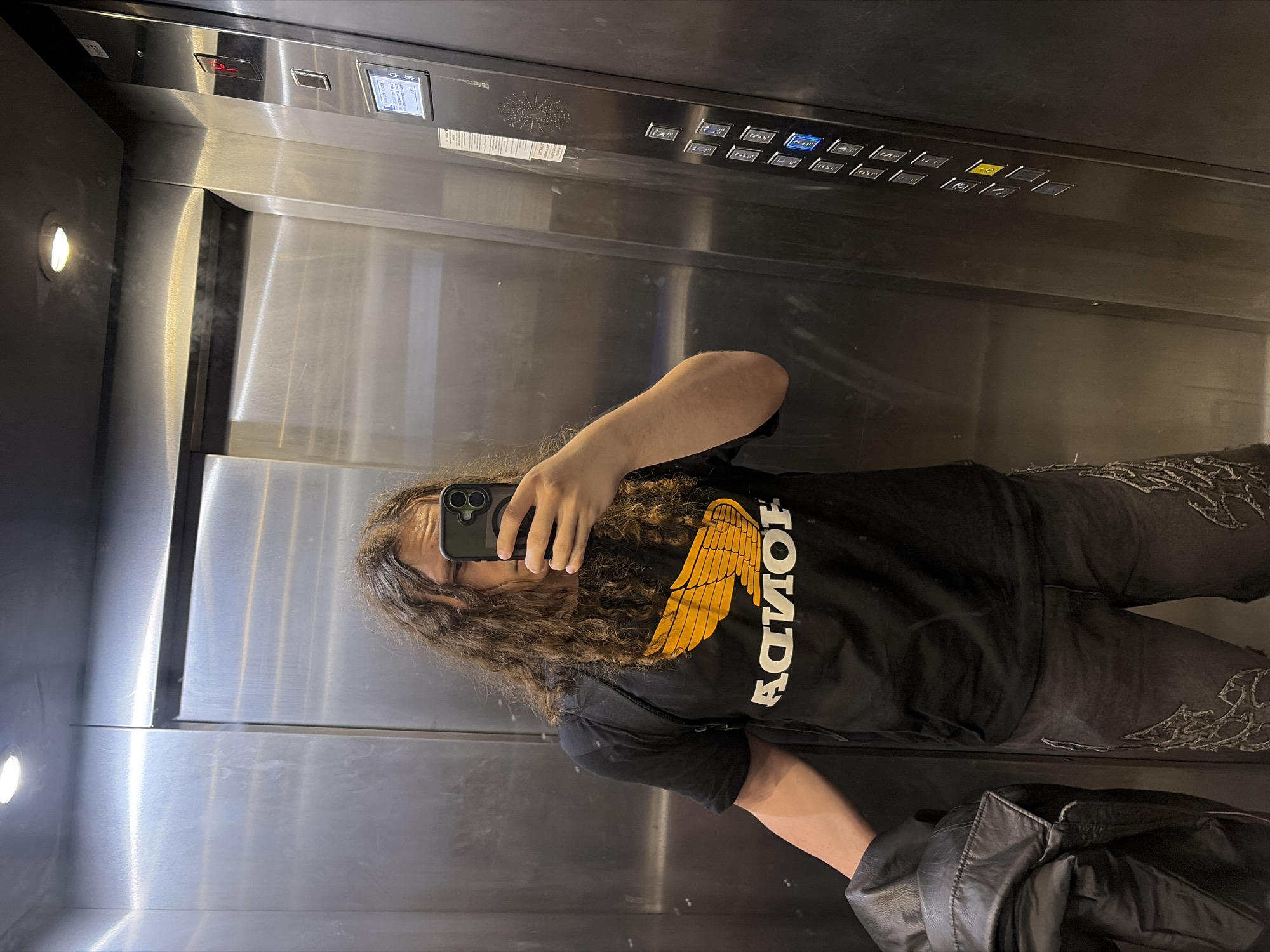A person is captured in a reflection selfie inside a modern elevator with brushed stainless steel walls. The individual, seen from the waist up, has long, curly dark hair and is wearing glasses. They are holding a dark-colored smartphone to take the picture, partially obscuring their face.
The person wears a black t-shirt featuring a large golden-orange graphic of wings and the word "HONDA" written in white (appearing reversed due to the reflection). They are also wearing dark-colored jeans with intricate white or light-toned patterns embroidered on the sides of the legs. A black leather jacket or bag is visible in the lower right corner of the frame.
The elevator interior is highly reflective, showcasing the metallic surfaces. On the control panel, numerous square buttons are visible, some illuminated with blue or yellow lights, likely indicating floor selections or functions. A speaker grill is present on the panel. A digital display near the top right of the panel shows what appears to be "4F" (indicating the 4th floor) along with an upward-pointing arrow and some Asian characters, possibly Chinese or Korean. Another display shows a red light, possibly an indicator for an alarm or emergency. Two circular lights illuminate the left wall of the elevator.
The image appears to be taken during the day or in an artificially lit indoor environment, given the bright interior lighting. No specific weather or time of day cues are discernible. The setting is an elevator, consistent with the provided context of Plovdiv, Bulgaria, though no external location markers for the city are present.
