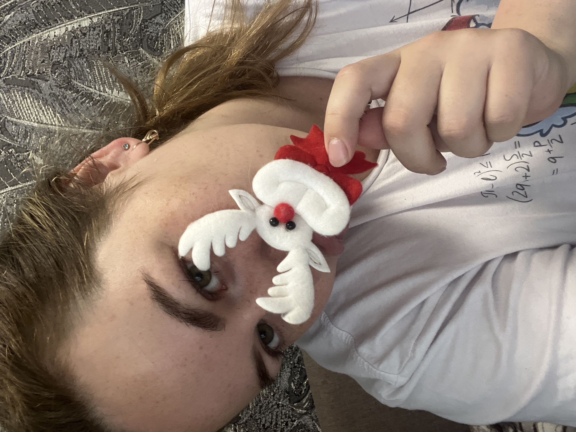 A person, likely female, is depicted lying down indoors in Chaikovsky, Russia, with their head and upper torso angled approximately 45 degrees clockwise. They have fair skin with freckles visible on their face and chest, medium-brown hair, and dark, well-defined eyebrows. Their eyes, appearing greenish-brown, are open and directed towards the camera.
The individual is wearing a plain white t-shirt, which is adorned with mathematical equations printed in black. Partially visible equations include "π - t^2 ≤", "(2q + 2) 5/2 = P", and "9 + 2 =". In their right hand, they hold a small, white felt reindeer figurine. The reindeer features a red nose, small black eyes, white antlers, and a miniature red Santa hat.
Visible details include a small stud earring in the person's left earlobe and a gold-colored piercing or cuff higher on the helix. The background consists of a dark, textured fabric with a subtle pattern, suggesting the person is resting on a bed or couch. The overall activity appears to be a casual pose, possibly taken playfully with the festive ornament. The lighting indicates an indoor setting, with no specific time of day or external weather conditions discernible.
