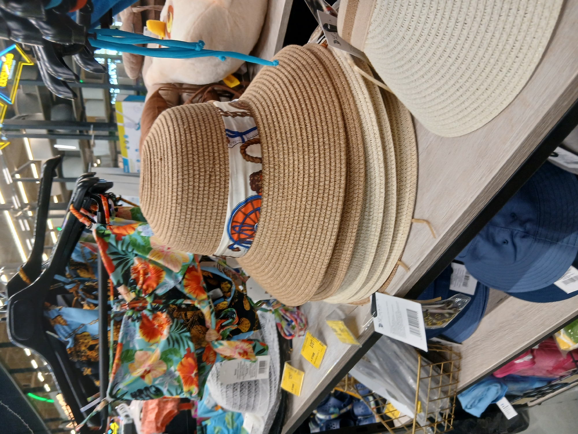The image shows a retail display of summer clothing and accessories, likely in a shop in Chabany, Ukraine. In the foreground, a stack of woven straw hats is displayed on a shelf. The top hat is decorated with a patterned ribbon featuring nautical motifs. Behind the hats, a white wire basket containing plastic-wrapped items is visible. Further back, on a higher shelf, floral-patterned swimwear is hanging on hangers. The background shows shelving units with various goods and lighting fixtures, typical of a retail environment. The overall scene depicts a well-stocked store with a focus on seasonal fashion items.
