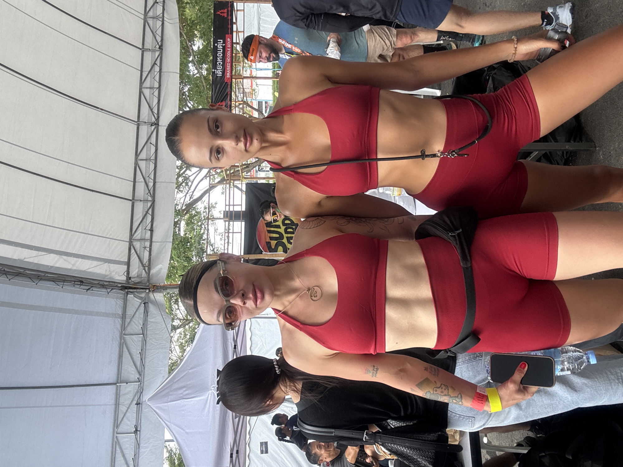 Two young women wearing matching red workout outfits stand side-by-side in a public setting. One woman wears sunglasses and has visible tattoos on her arm. They appear to be at an outdoor event in บ้านสำนักตะแบก, Thailand, indicated by a sign in Thai script in the background. There are other people around, some sitting and some walking. The location seems to be under a temporary shelter, likely a tent, as indicated by the supports and fabric visible in the background. One of the women is holding a phone and a water bottle.