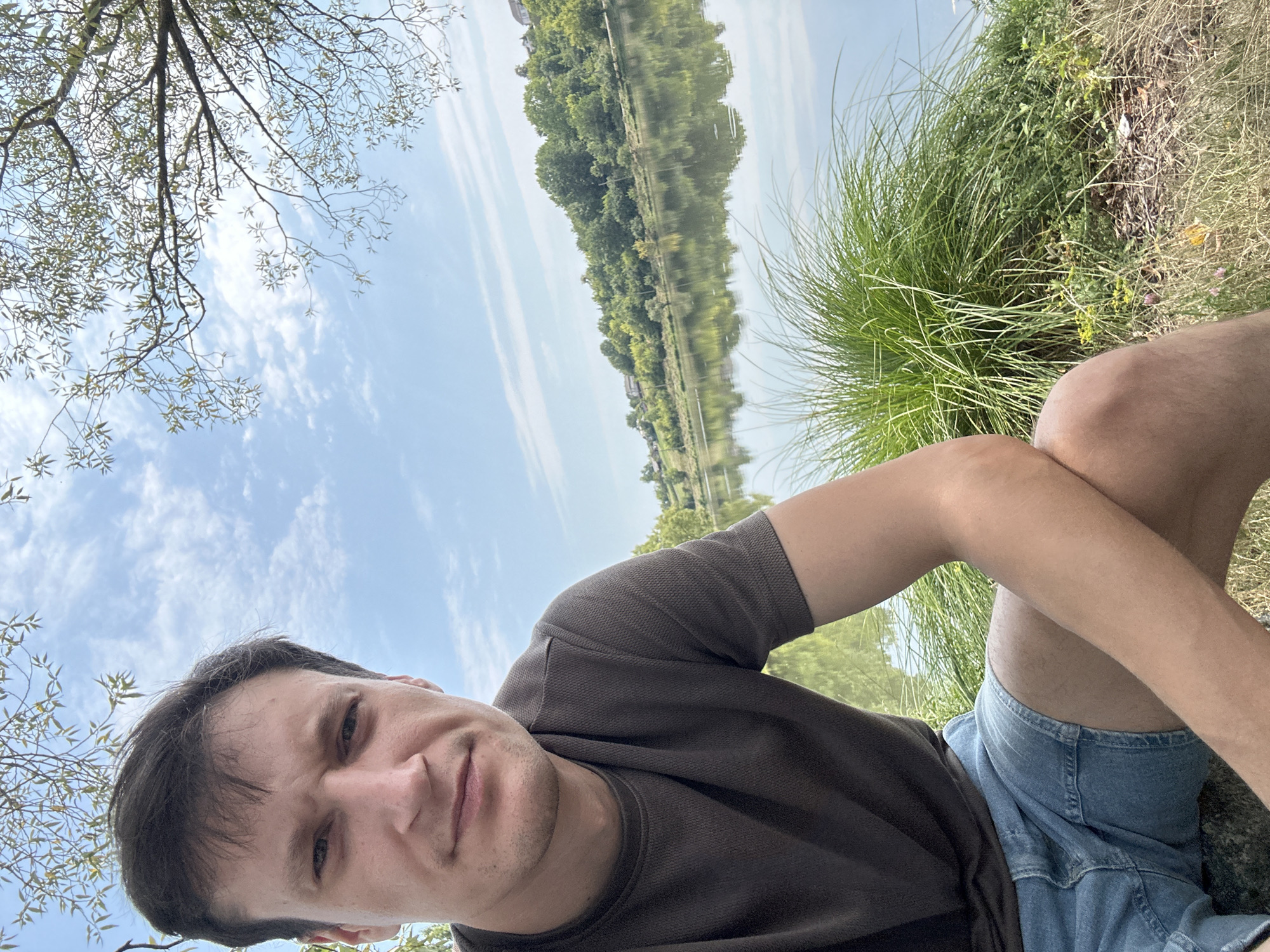 A medium shot selfie shows a young adult male in the foreground sitting outdoors by a body of water, possibly a lake or river. He is looking directly at the camera with a neutral expression. He is wearing a dark brown textured t-shirt and light blue denim shorts. His left arm is resting on his leg. Behind him, lush green grass and reeds grow along the water's edge. The water reflects the blue sky with scattered white clouds, and a tree-lined bank with houses is visible in the distance. The scene appears to be set in Makiv, Ukraine, suggesting a natural outdoor location within or near the city.