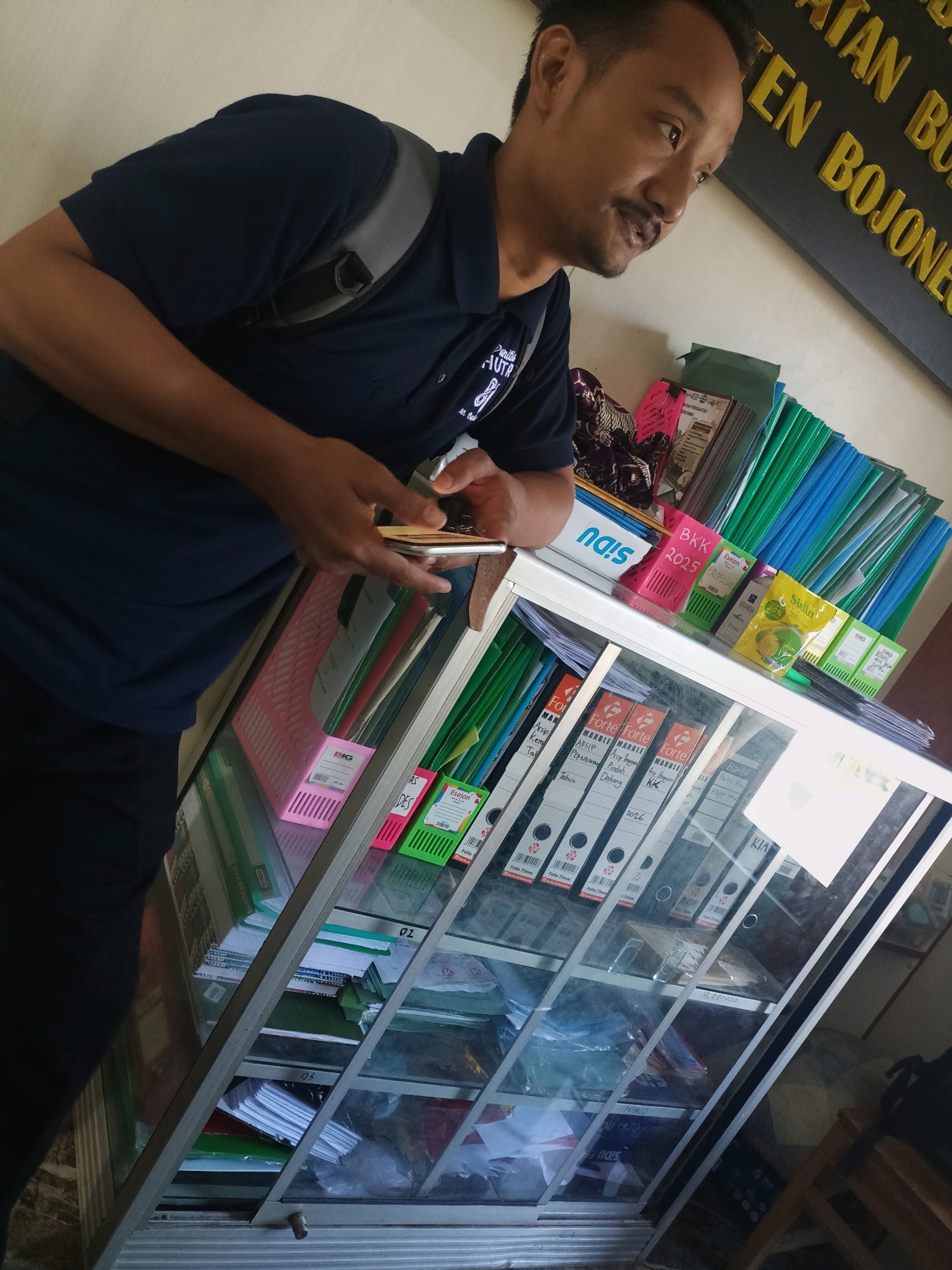 A man with dark hair, a mustache, and light brown skin is holding a smartphone in his right hand and looking down and to his right. He is wearing a dark blue polo shirt and a dark backpack. The man is standing in front of a glass-fronted display cabinet filled with files and folders. The cabinet has a white frame and is filled with various colored folders, binders, and stacks of paper. Behind the man, a dark sign with yellow lettering is visible on a light-colored wall. The lettering on the sign appears to be in Indonesian. The overall scene suggests an indoor office or storage environment. Based on the sign, the location is identified as Sumber Bendo, Indonesia.