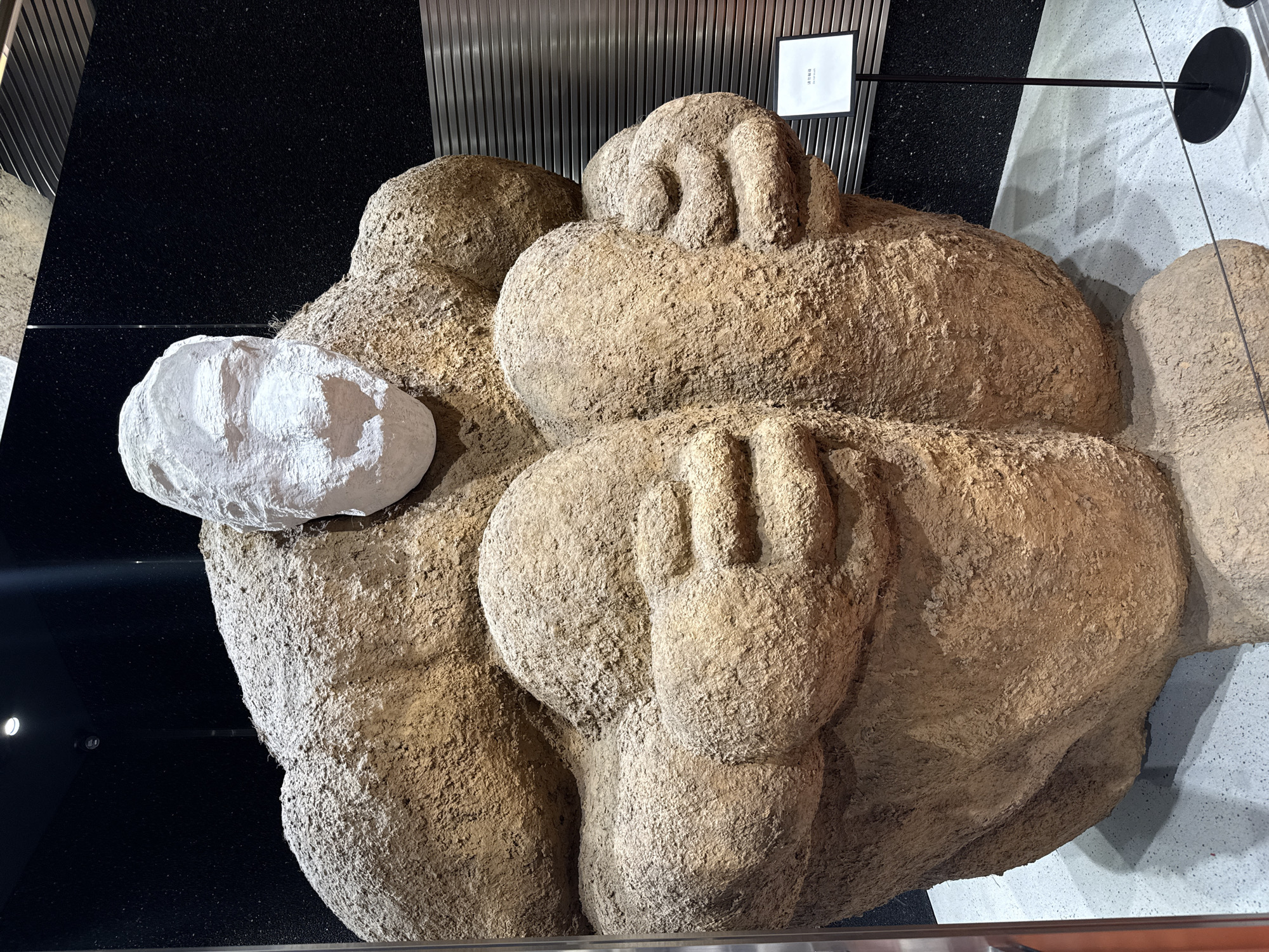 The image shows a large sculpture of entwined figures, possibly human-like, with a rough, earthen texture. The sculpture appears to be indoors, located near a wall with a dark background on one side and a lighter, potentially marble-like surface on the other. A white, rock-like object is positioned near the figures. There's also a sign with text written in a non-English language. While the location is stated to be in the Tianhe District, China, this cannot be confirmed from visual data alone.