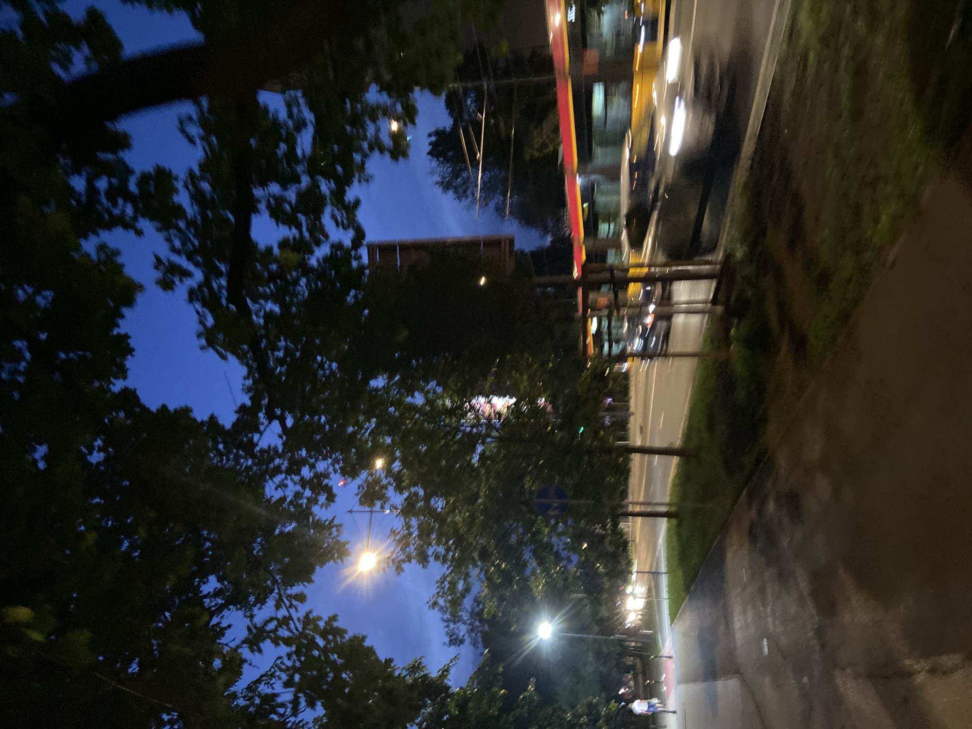 The image depicts a street scene at dusk in Warsaw, Poland. A sidewalk and a road are visible, with trees lining the street. A person is walking on the sidewalk, and a few cars and a tram are on the road, captured with motion blur. Streetlights are illuminating the scene. The sky is a deep blue, indicating it is either early evening or late night.
