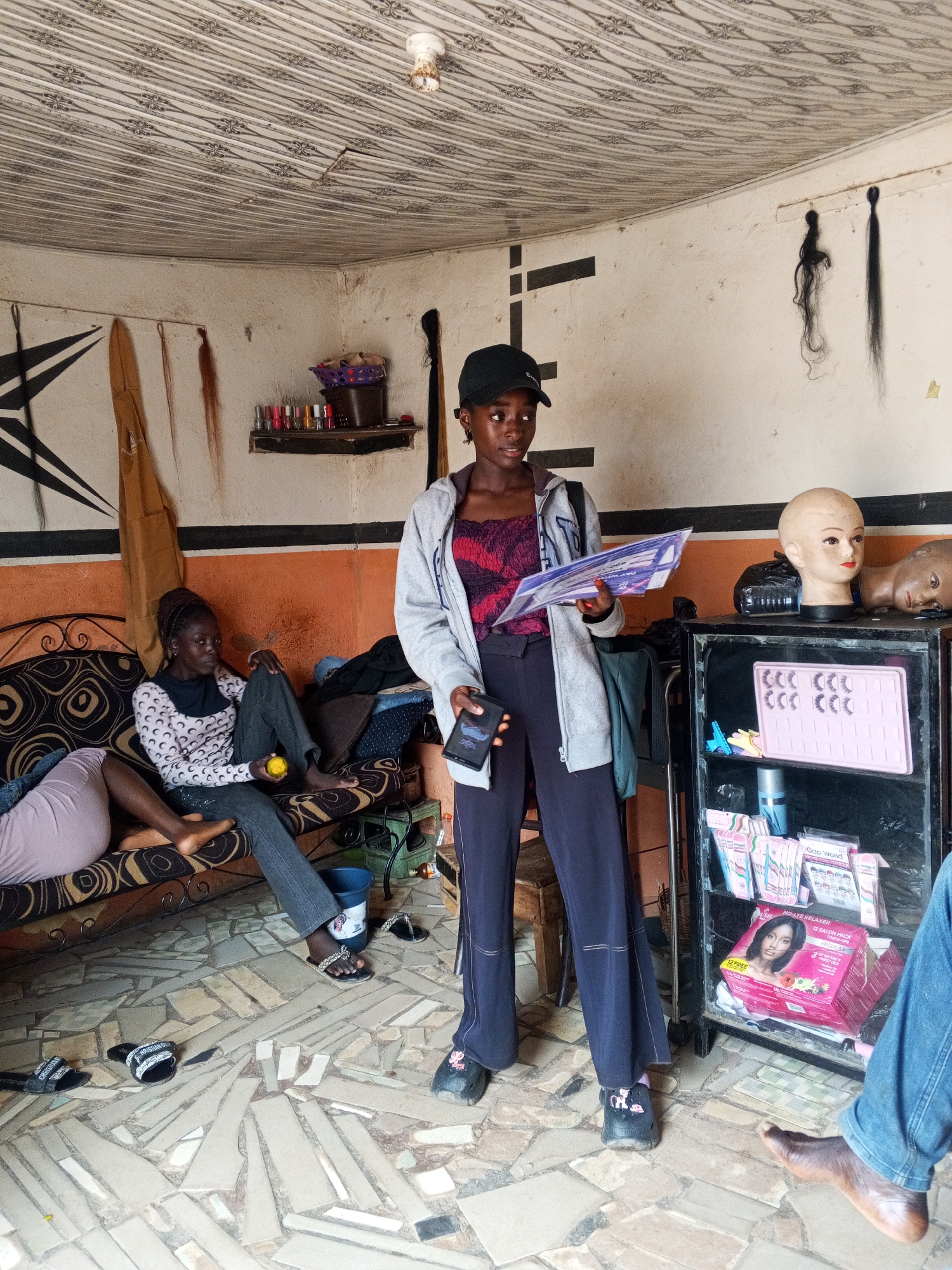 The scene depicts the interior of a salon, possibly located in Gwong, Nigeria. A young woman wearing a black cap, gray hoodie, maroon top, and dark pants stands in the foreground, holding a pamphlet in her right hand and a phone in her left. A black cabinet filled with hair products, including a pink box with a woman's image on it, stands to her right. Two mannequin heads sit atop the cabinet.
A woman is seated on a brown patterned couch in the background, holding a yellow object. Another person is partially visible on the same couch. A shelf with cosmetics and a purple basket is mounted on the wall above the couch. The walls are painted with orange and white stripes and decorated with geometric patterns and hanging hair extensions. The floor is tiled with irregularly shaped tiles. Various items, including shoes and a bucket, are scattered on the floor. The ceiling is patterned. A person wearing blue jeans is partially visible in the lower right corner of the image.