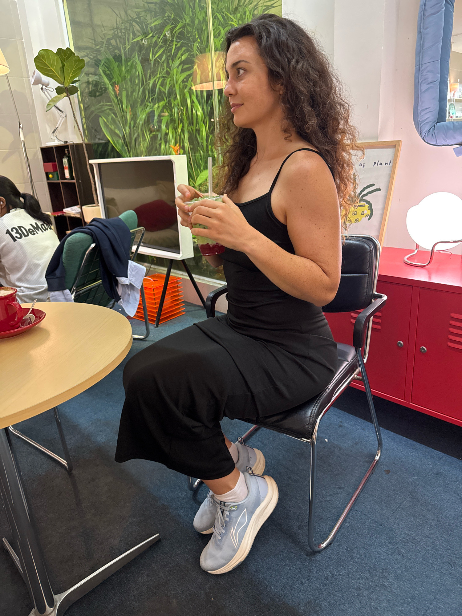 A young woman with curly brown hair is seated in a black office chair at a cafe. She is wearing a black sleeveless dress and light blue athletic shoes with white socks. She holds a clear plastic cup with a green beverage and a straw. To her left, a round, light wood table has a red coffee cup and saucer on it. Behind her, a large window displays lush green foliage, suggesting an indoor setting with an outdoor view. In the background, other cafe elements are visible, including shelves with items, a monitor, and a red cabinet with lockers. The floor is covered with blue carpeting. The woman's gaze is directed to her right, away from the camera.