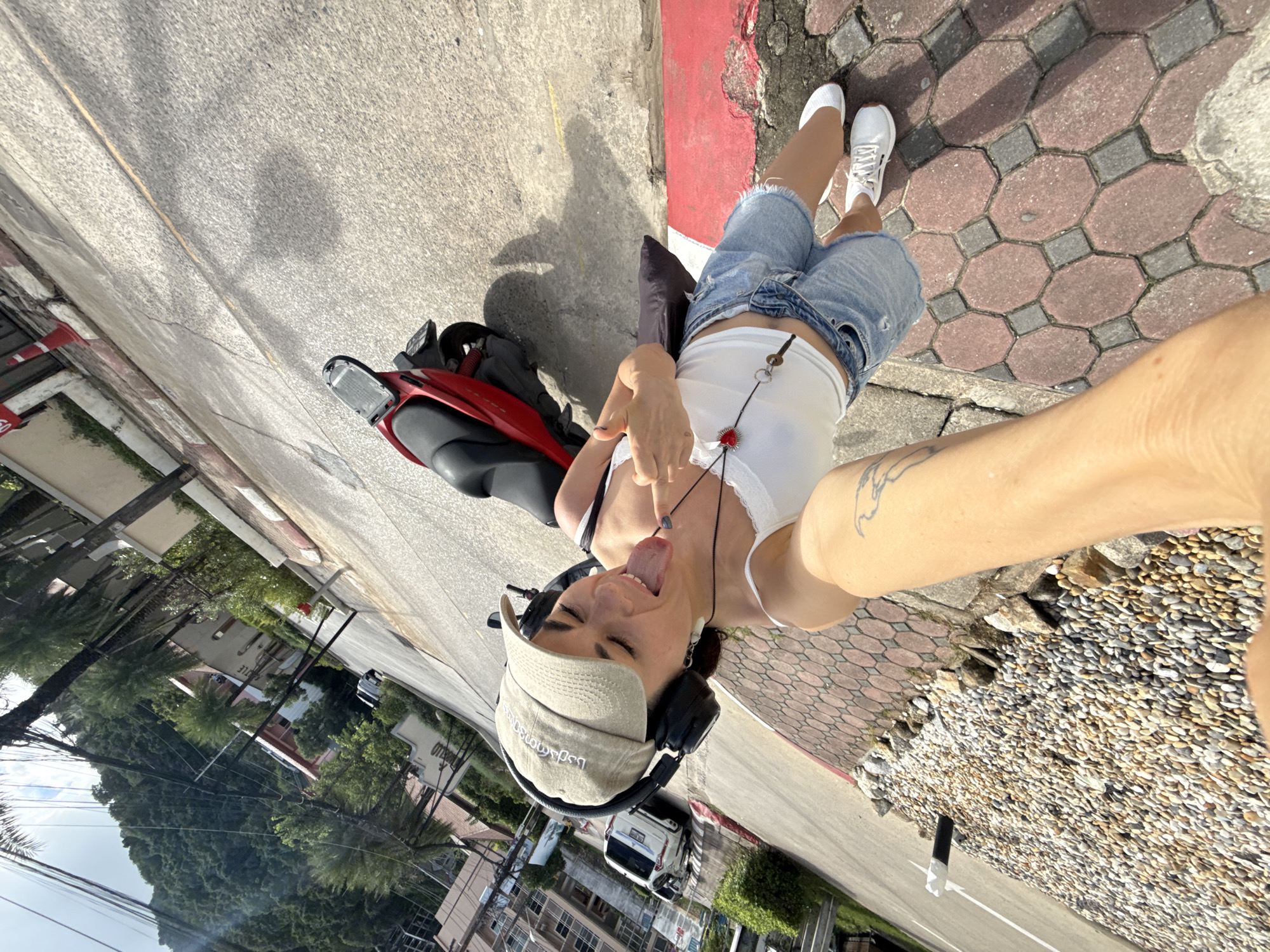 A woman is taking a selfie while standing next to a red motorcycle on a street in Karon, Thailand. She is wearing a white tank top, denim shorts, a tan baseball cap, and headphones. She is sticking her tongue out and giving a middle finger to the camera. The street is paved with asphalt and has a red and white curb. There are buildings and lush green hills in the background.