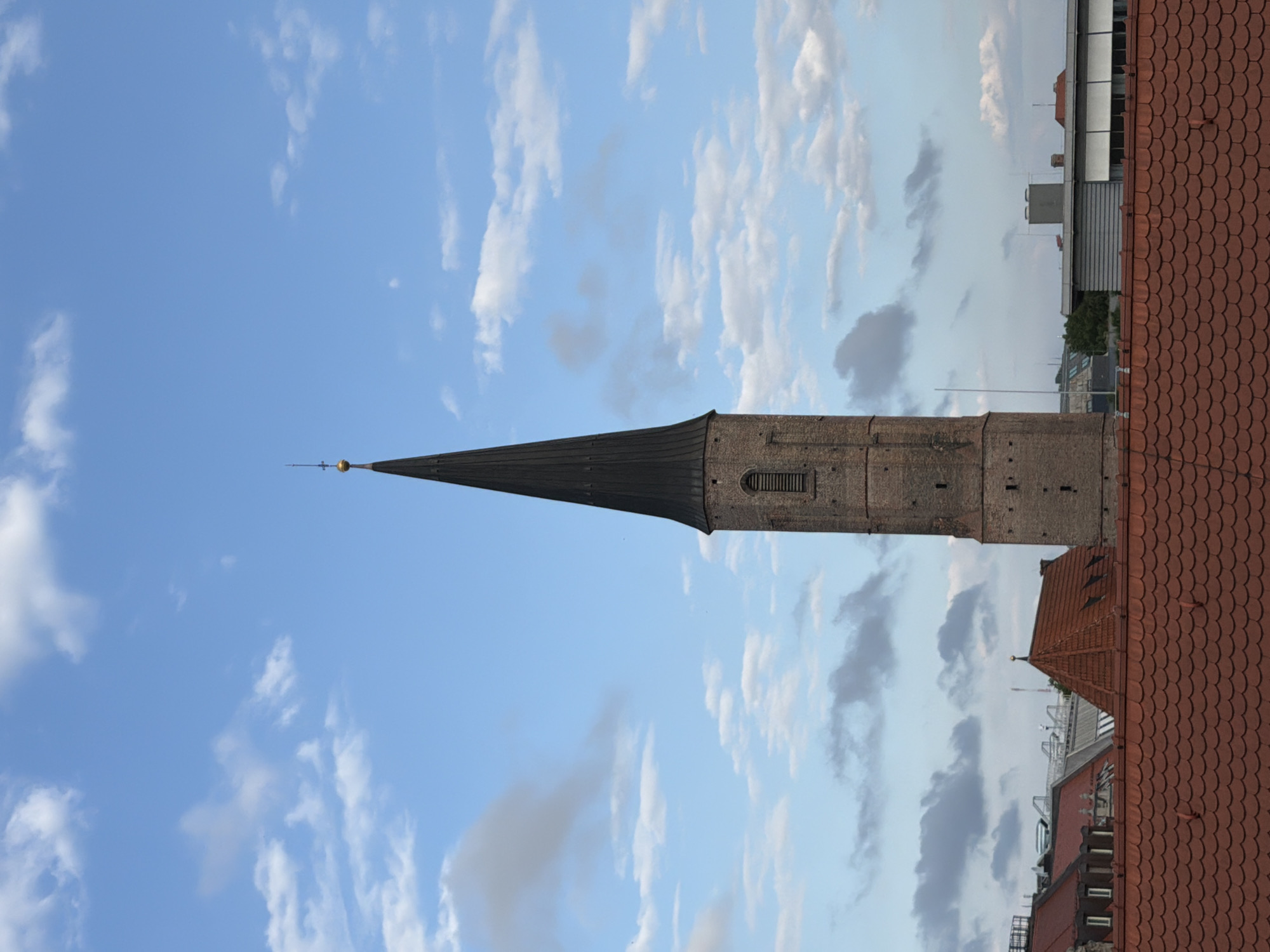 The image shows a close-up view of the spire of St. Peter's Church in Munich, Germany, against a bright blue sky with scattered white clouds. The spire, constructed of brick and topped with a dark conical roof, dominates the frame. Below the spire, red-tiled rooftops of other buildings are visible, characteristic of the city's architecture. The overall scene depicts an urban landscape under a partly cloudy sky.