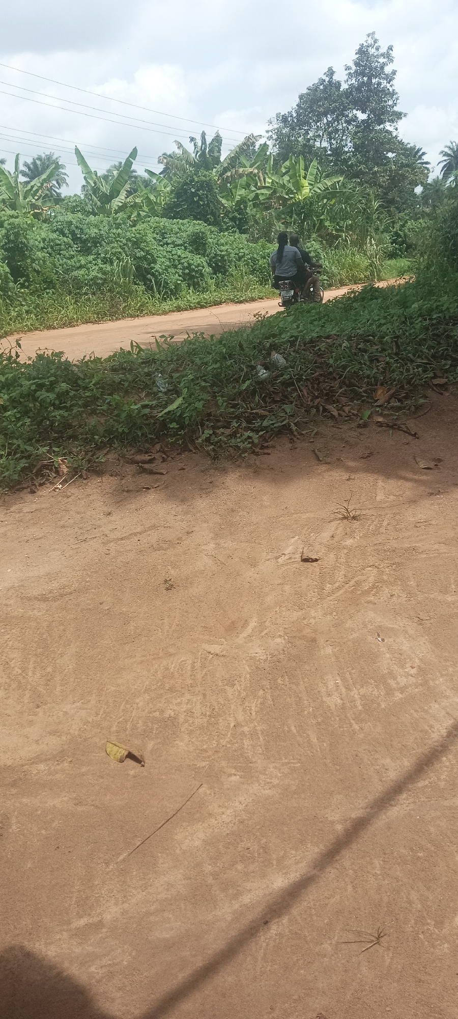 A medium shot depicts a dirt road in Amauzu, Nigeria, with lush green vegetation bordering both sides. A motorcycle carrying two people is visible on the road in the distance. Overhead power lines cross the scene. The foreground shows the dirt road's texture, with scattered leaves and a shadow cast by an unseen object. The sky is overcast.