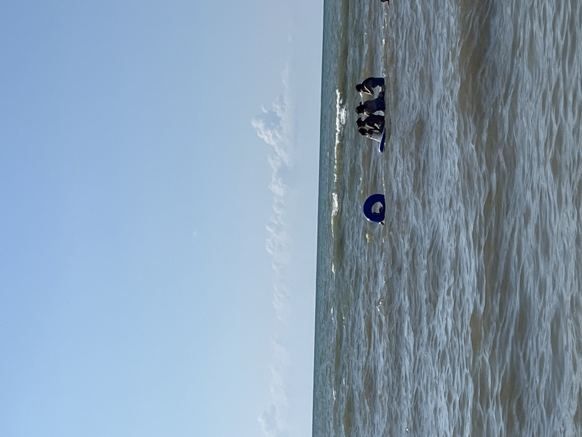 In the waters of Ureki, Georgia, a group of four individuals are seated on a blue flotation device. They are positioned in the sea, with waves gently lapping around them. To the left of the group, a blue inflatable ring floats in the water. The sky above is clear blue with sparse, wispy clouds. The overall scene depicts a relaxed day at the beach.