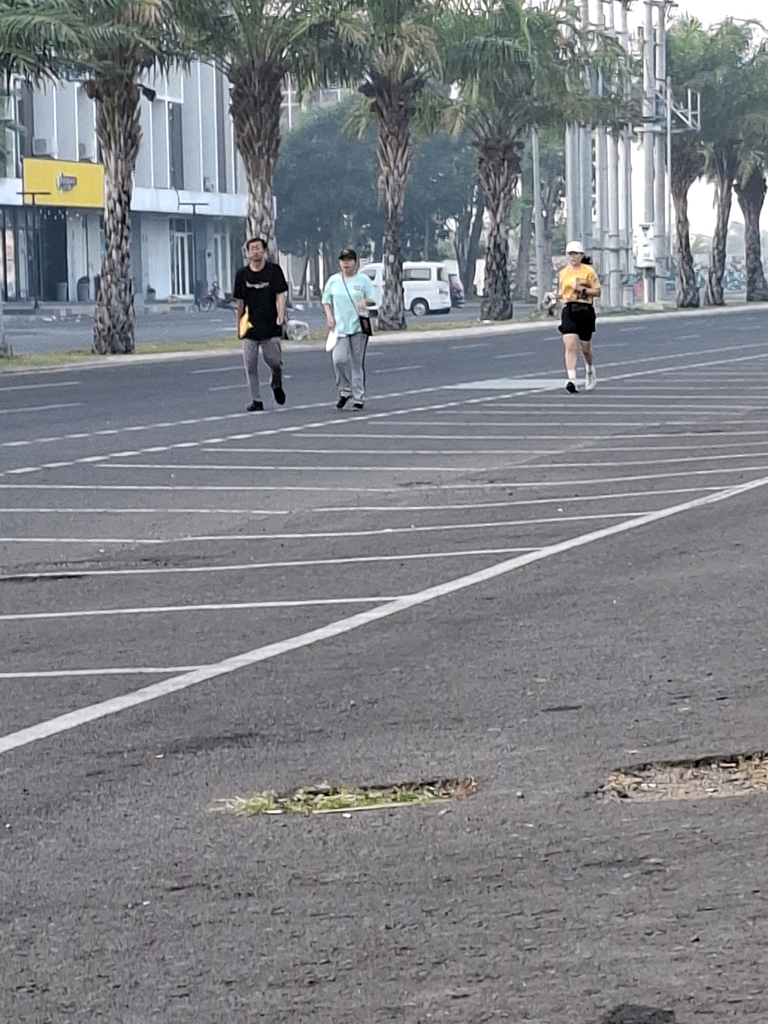 Three people are walking or jogging on a paved surface with white lines, possibly a running track or a road with marked lanes. In the background, there are palm trees, buildings with glass windows, and utility poles. A white van is parked on the left side of the frame. The lighting suggests it is daytime. The image appears to have been taken in Surabaya, Indonesia, based on the surrounding environment.