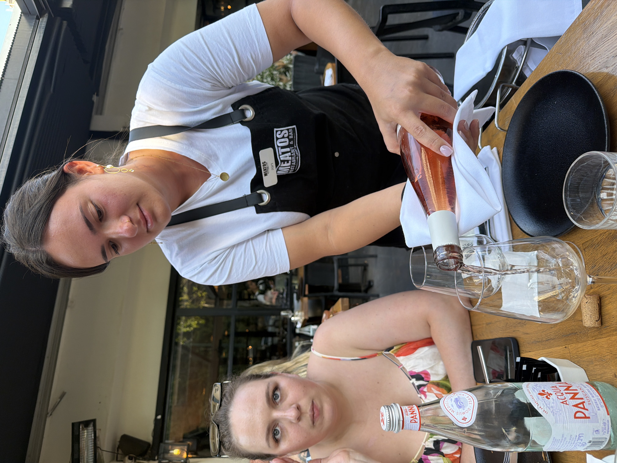 A woman wearing a white t-shirt and a black apron with the logo "MEATOS RESTAURANT & BAR" is pouring rosé wine from a clear bottle into a wine glass. She is standing at a wooden table, and another woman with blue eyes and blonde hair, wearing a colorful dress, is seated at the table. To the left of the seated woman is a bottle of Acqua Panna water. The background shows an outdoor dining area with tables and chairs. The scene appears to be in Tel-Aviv, Israel.