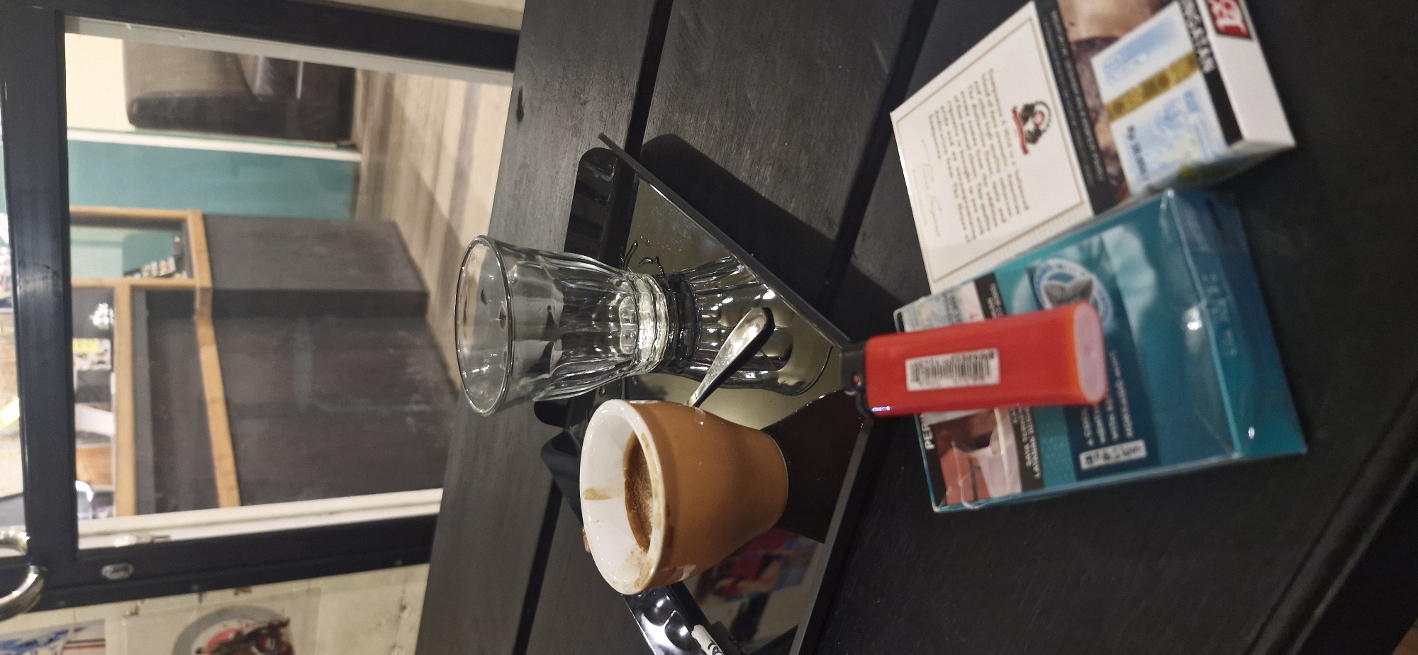 A close-up, low-angle shot captures a dark wooden table in what appears to be a cafe or bar in Rantau, Indonesia. In the foreground, a teal-colored cigarette pack rests beside a red lighter and a white packet with blue text. Further back, a small brown ceramic cup filled with dark liquid, likely espresso, sits on a reflective black tray next to a clear, fluted glass. The background is blurred, showing glimpses of a counter area and seating through a glass partition.
