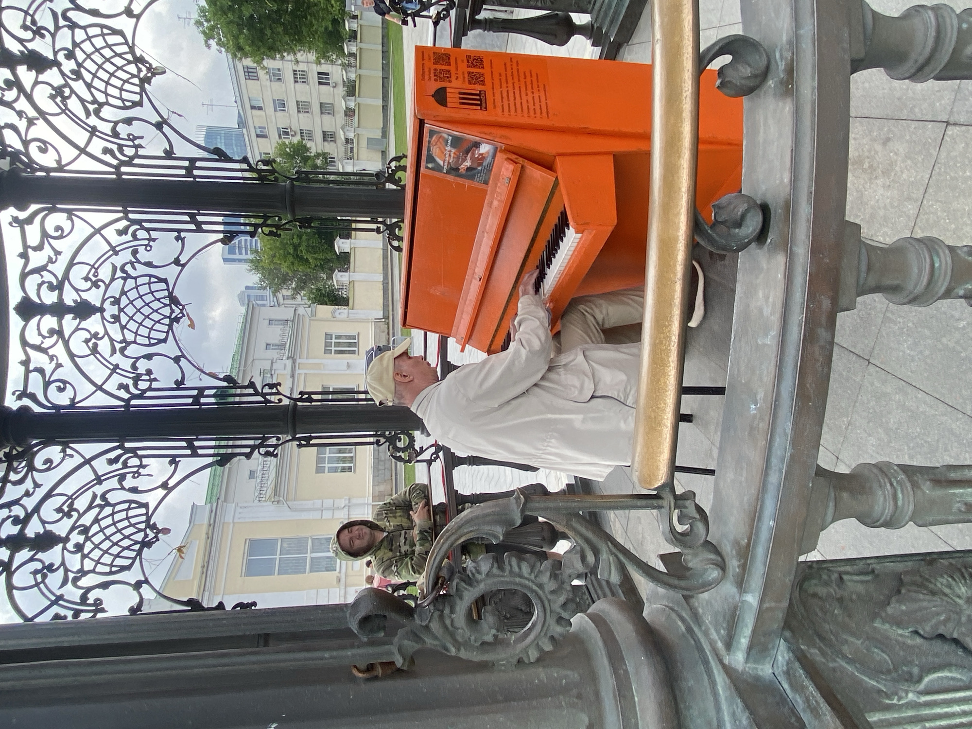 An older Caucasian male wearing a light-colored jacket and a tan baseball cap plays an orange upright piano. He is seated and his hands are on the keys. The piano is positioned in an open-air gazebo with ornate metalwork. A younger Caucasian male wearing a camouflage hoodie stands to the left of the piano player, leaning against a railing and looking towards the piano player. The background shows a city environment with buildings and trees. The gazebo appears to be made of dark metal with intricate scrollwork. The ground is paved with light gray square tiles. The image is taken from a slightly low angle, looking up at the gazebo and the subjects. The overall lighting suggests an overcast day.