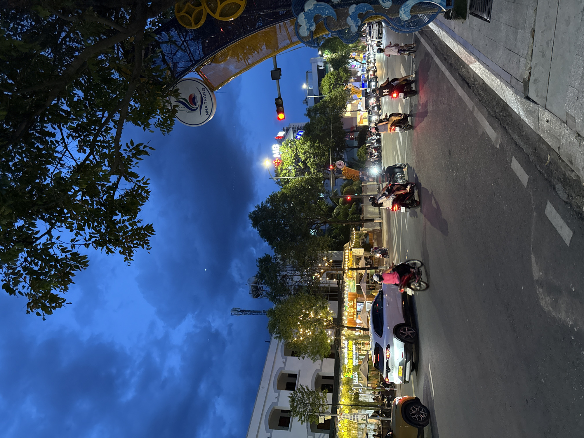 The image depicts a street scene in Nha Trang, Vietnam, at dusk. The sky is a deep, bruised blue with scattered clouds, and a sliver of the moon is visible. A large tree with dark green foliage arches over the top right of the frame, partially obscuring the sky.
Below the tree, a circular sign with the words "NHA TRANG" is visible, suggesting the location. A decorative archway with blue wave-like motifs and yellow circular elements stands to the right.
The street itself is a busy thoroughfare with several lanes of traffic. Motorcycles are the dominant mode of transportation, with many individuals riding them. A white SUV is stopped in the middle lane, and a yellow car is partially visible on the left edge of the frame. Pedestrians are present on the sidewalk to the right.
Buildings line the street, some of which are illuminated with strings of lights. One building on the left appears to be a commercial establishment with awnings and umbrellas set up outside. A traffic light is visible, showing a red signal. The overall atmosphere suggests a vibrant urban environment transitioning into evening.