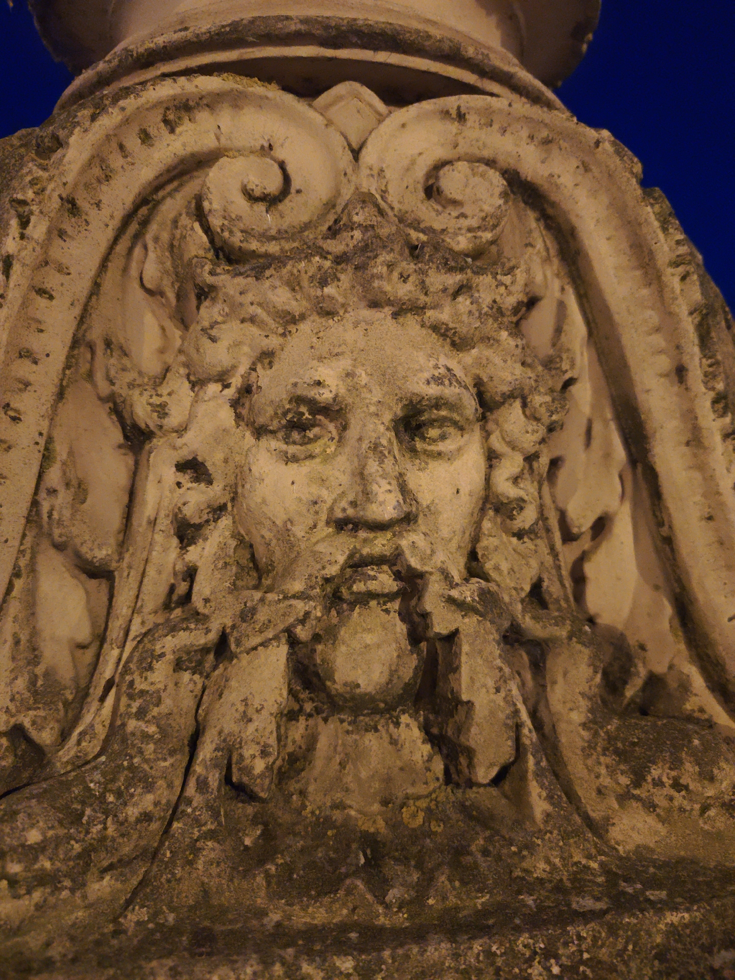 The image displays a close-up, low-angle shot of an ornate architectural detail, possibly a grotesque or gargoyle, carved from stone. The carving depicts a weathered, human-like face with a beard and curly hair, framed by decorative scrollwork. The stone is textured and appears aged, with patches of moss or grime. The background is a deep blue, suggesting it is nighttime. This architectural element is located on the exterior of the Schwerin Castle in Schwerin, Germany.