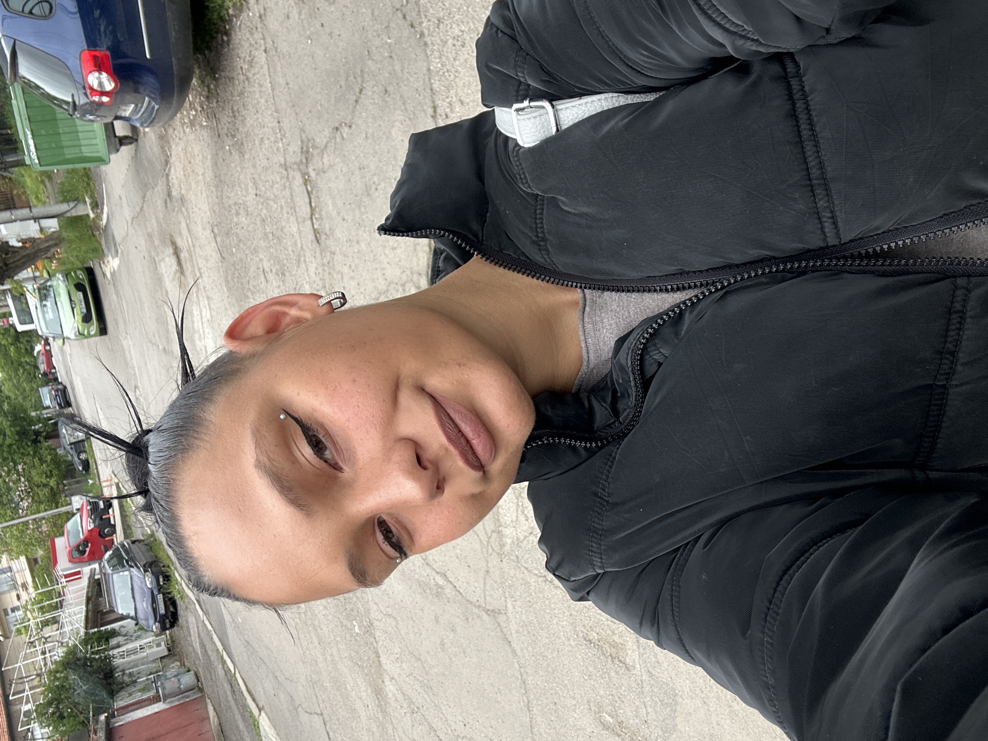 A medium-shot selfie depicts a person with fair skin, brown eyes, and dark hair pulled back into a messy bun, with a small eyebrow piercing. They are wearing a black puffer jacket with a gray shirt underneath. A white strap is visible across their upper body.
The background reveals an outdoor urban setting. A concrete road occupies the lower portion of the frame. Parked cars of various colors (blue, green, red, black) are visible along the sides of the road. Buildings with weathered facades and metal fencing are also present.
The location is presumed to be in Sofia, Bulgaria, based on visual characteristics such as architectural styles, vehicle registration plates, and general urban landscape, which are common in Bulgarian cities.