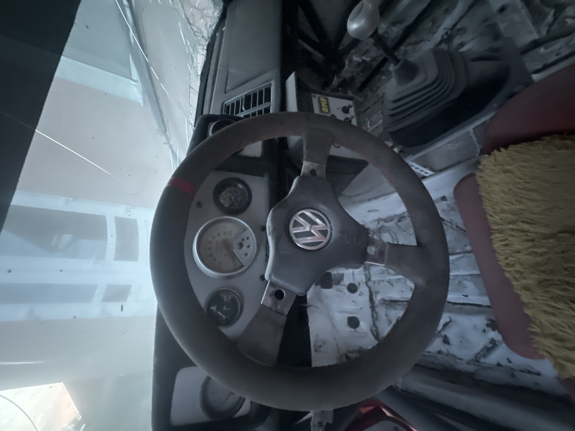 The image shows the interior of a car, likely a vintage Volkswagen, as evidenced by the logo on the steering wheel. The car appears to be in a state of disrepair, with a cracked windshield, exposed metal interior, and a gear shift lever. The dashboard has several gauges, including a speedometer and a tachometer. There is a red seat partially visible in the foreground, covered with a yellow, shaggy material. The overall impression is of an abandoned or project car. The environment suggests it is located in an urban setting, possibly a workshop or storage area.