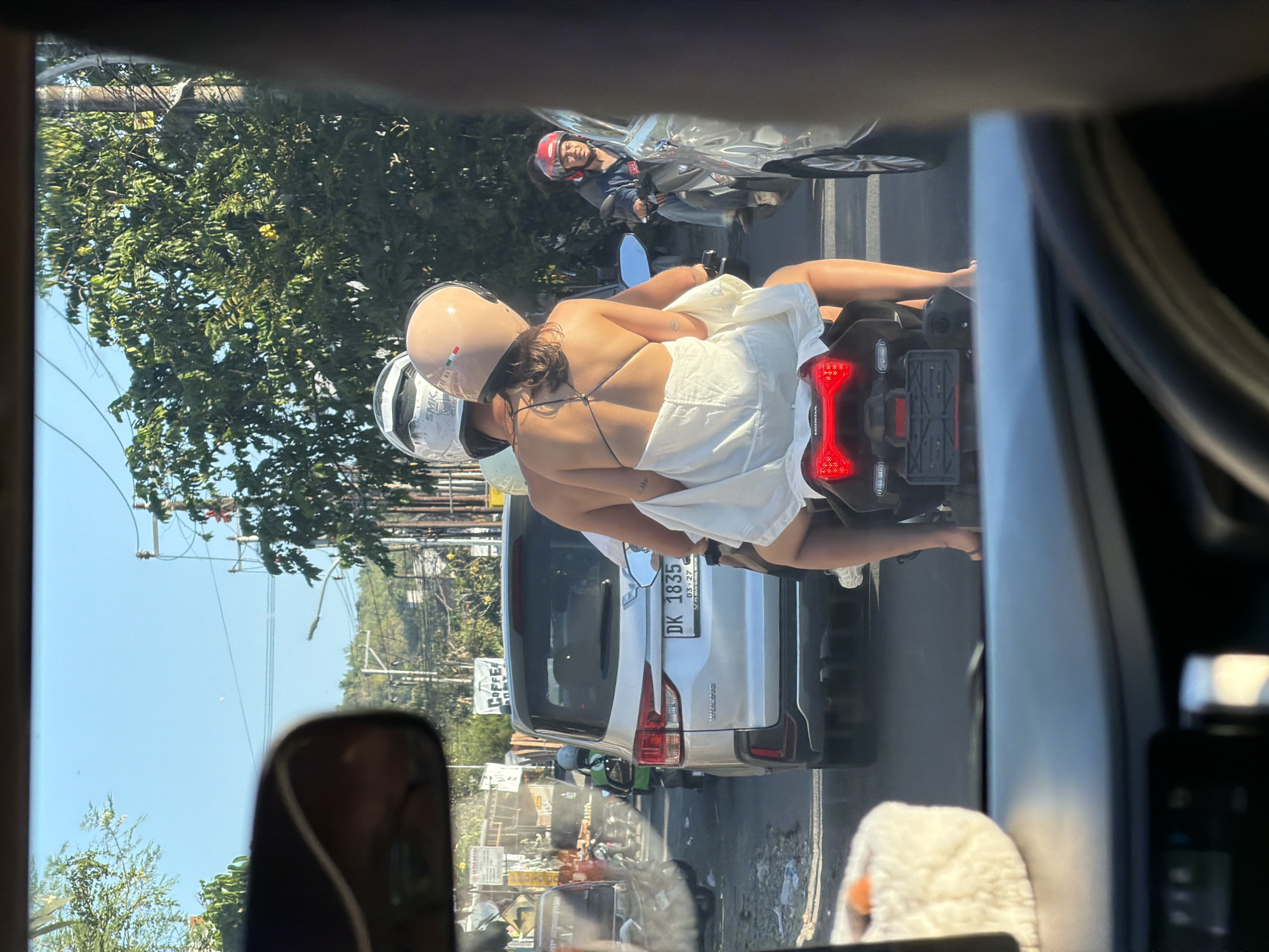 This image captures a snapshot of a bustling street scene in Kuta Selatan, Indonesia, likely during the day given the bright sunlight. The perspective is from inside a vehicle, looking out through the windshield and side mirror.
The central focus is on a motorcycle carrying two passengers, a man and a woman. The woman, in the front, is wearing a white dress that is draped over her. She has on a light-colored helmet. The man behind her is also wearing a helmet and appears to be driving. They are both looking ahead. Another motorcycle is visible behind them, with its rider wearing a red and black helmet. The motorcycles are navigating through traffic, as indicated by the presence of a white SUV in front of them with the license plate "DK 1835". Various shops and signs line the street, with lush green trees and power lines forming part of the background.
The overall atmosphere suggests a typical day on Indonesian roads, with traffic and pedestrians. The sunlight is strong, casting shadows and highlighting the vibrant colors of the environment. The image conveys a sense of movement and the daily life of a busy locality.