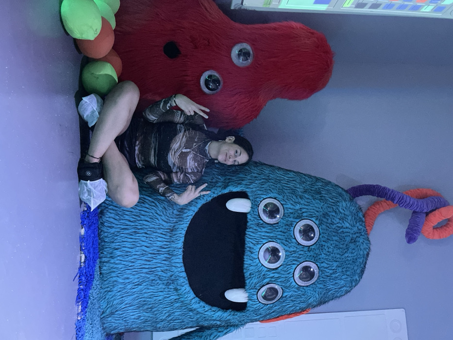 A woman lies sideways, propped between a fluffy blue monster with multiple eyes and a large, red, fuzzy blob, also with eyes. She wears a patterned top, black shorts, and shoe covers. She is flashing the peace sign with both hands. A string of orange, red, and green balls is visible, as well as purple and orange tendrils. The room is lit with a blue hue.