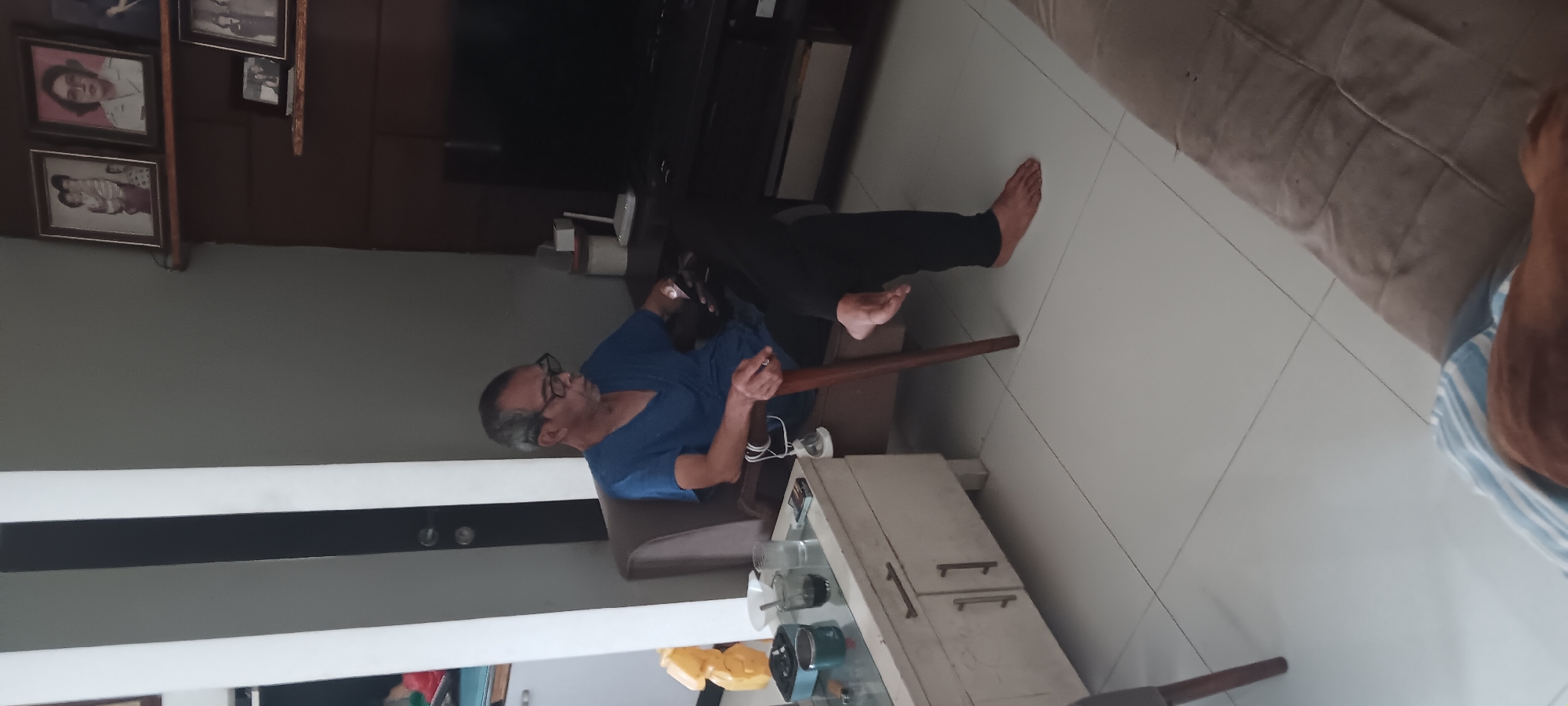 The image shows an elderly man wearing glasses and a blue t-shirt sitting in a chair. He is looking down at his phone, which he is holding in his hands. A wooden cane rests against the chair beside him. In front of the chair is a low white cabinet with two drawers. On the cabinet are several items, including a green cylindrical object, a white bowl, two glasses, and a pack of cigarettes. The man is wearing black pants and is barefoot. The floor is tiled with white squares. In the background, there is a dark brown entertainment center with a large black television screen. On shelves above the entertainment center are framed photographs. The room appears to be a living room in a home. The overall lighting is dim.