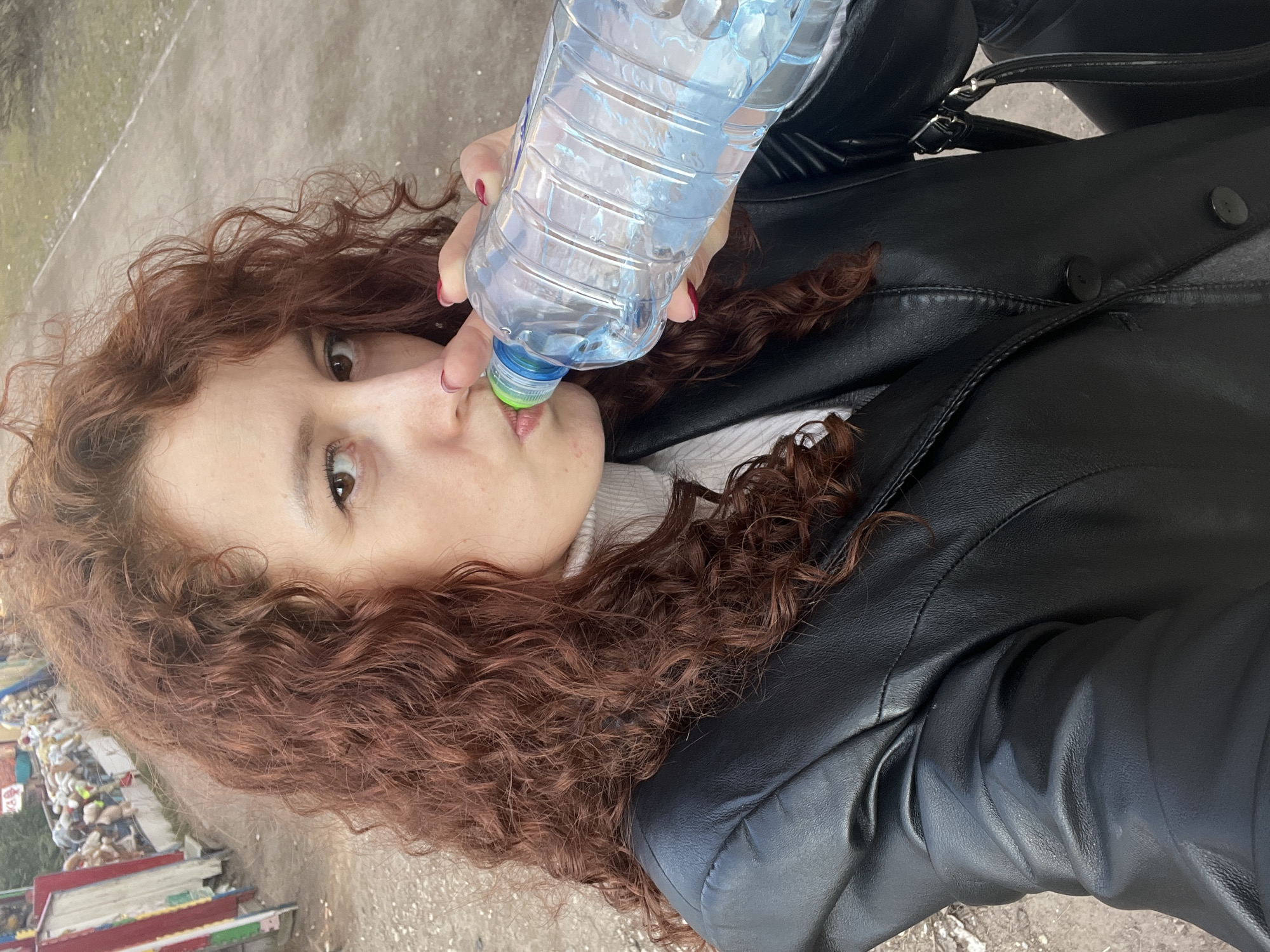 A woman with long, curly auburn hair is holding a plastic water bottle to her lips. She is wearing a black leather jacket over a white turtleneck sweater. The location appears to be a city street. In the background, there is what appears to be a pile of garbage or debris. It is not possible to confirm the specific location in Saint Petersburg, Russia from the image.