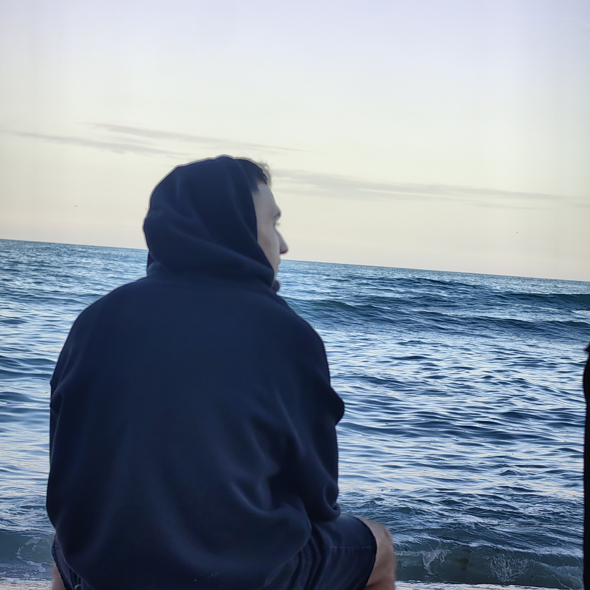 A person wearing a dark hoodie sits on a beach looking out at the sea. The person is facing away from the camera, and only the side of their face is visible. The sea is blue and choppy, and the sky is a pale blue with some clouds. The location may be a beach near the city of Varna, Bulgaria.