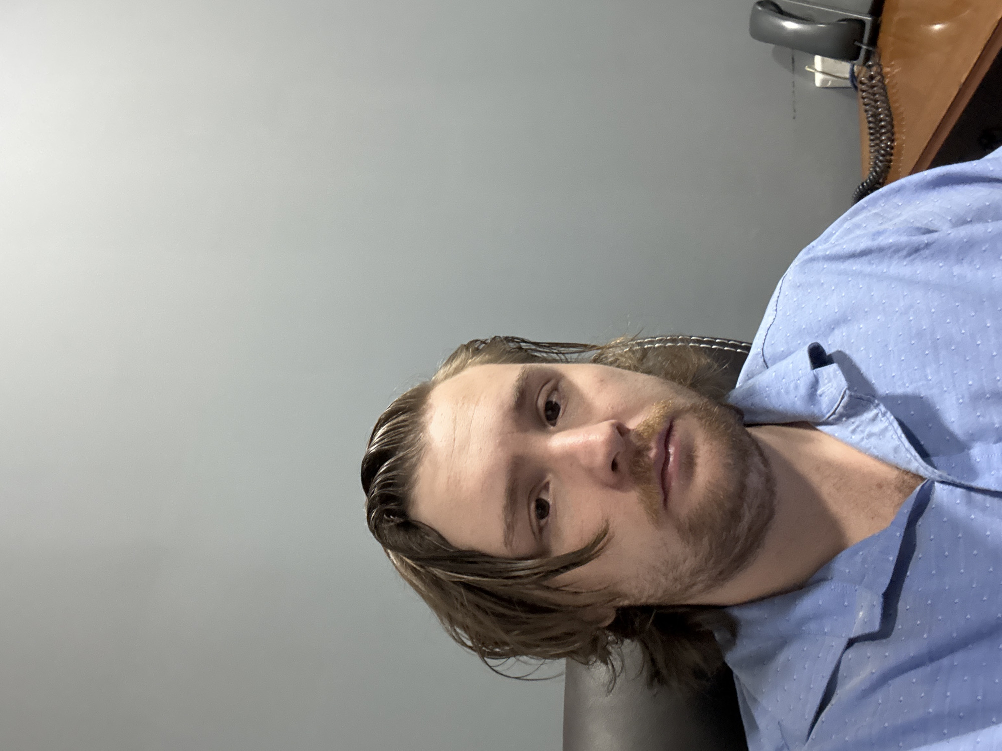 The image shows a man with shoulder-length brown hair, a light beard, and wearing a light blue button-down shirt. He is seated, possibly in a chair. The background is a light gray wall. A black telephone and wooden desk are visible in the upper right corner of the frame. There is no information to determine the location in Cuautitlán Izcalli, Mexico.