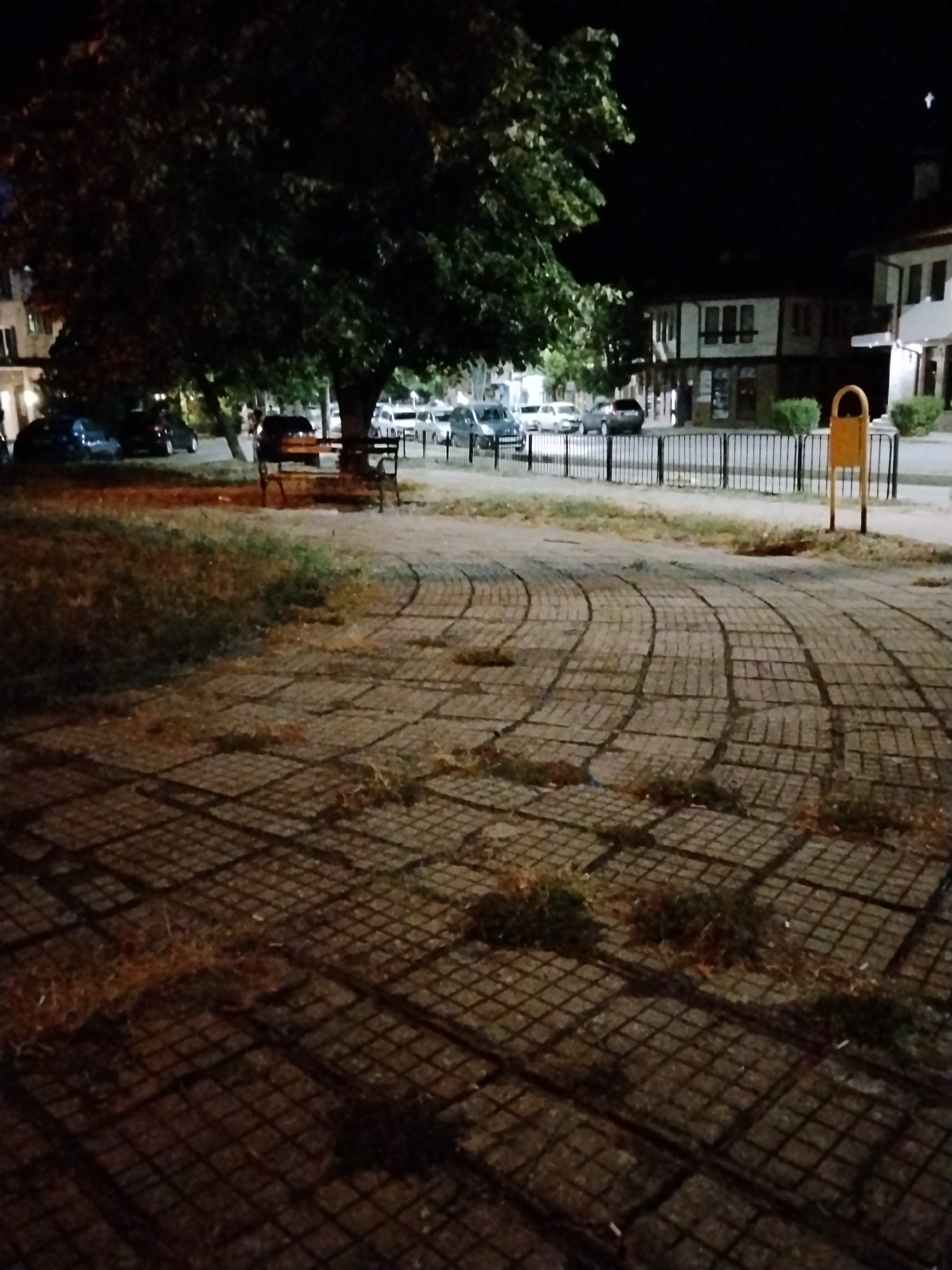 The image depicts a nighttime scene in Sliven, Bulgaria, likely a park or public square. A large tree with dark foliage dominates the left side of the frame, casting shadows. In the middle ground, a park bench is visible under the tree, with several cars parked behind it. A black metal fence runs horizontally across the scene, separating the paved area from the street where the cars are parked. Buildings with windows are visible in the background on the right. The foreground features a paved walkway made of square tiles arranged in a curved pattern, with patches of grass growing through the gaps. A yellow trash receptacle stands to the right of the fence. The lighting suggests artificial illumination from streetlights.