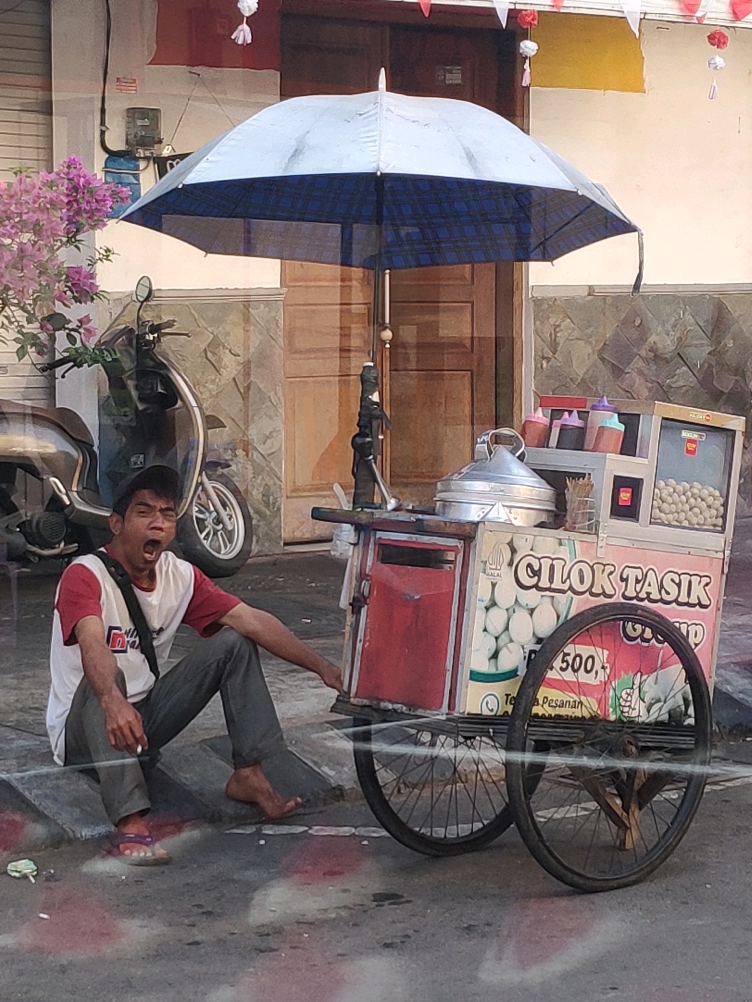 A man is sitting on the curb next to a food cart. The food cart has an umbrella over it. The man is wearing a red and white shirt and gray pants. He is yawning. The food cart is selling "Cilok Tasik". There are balls of food in a display case on the cart. A motorcycle is parked behind the man. The scene is on a street in Kebonagung, Indonesia.