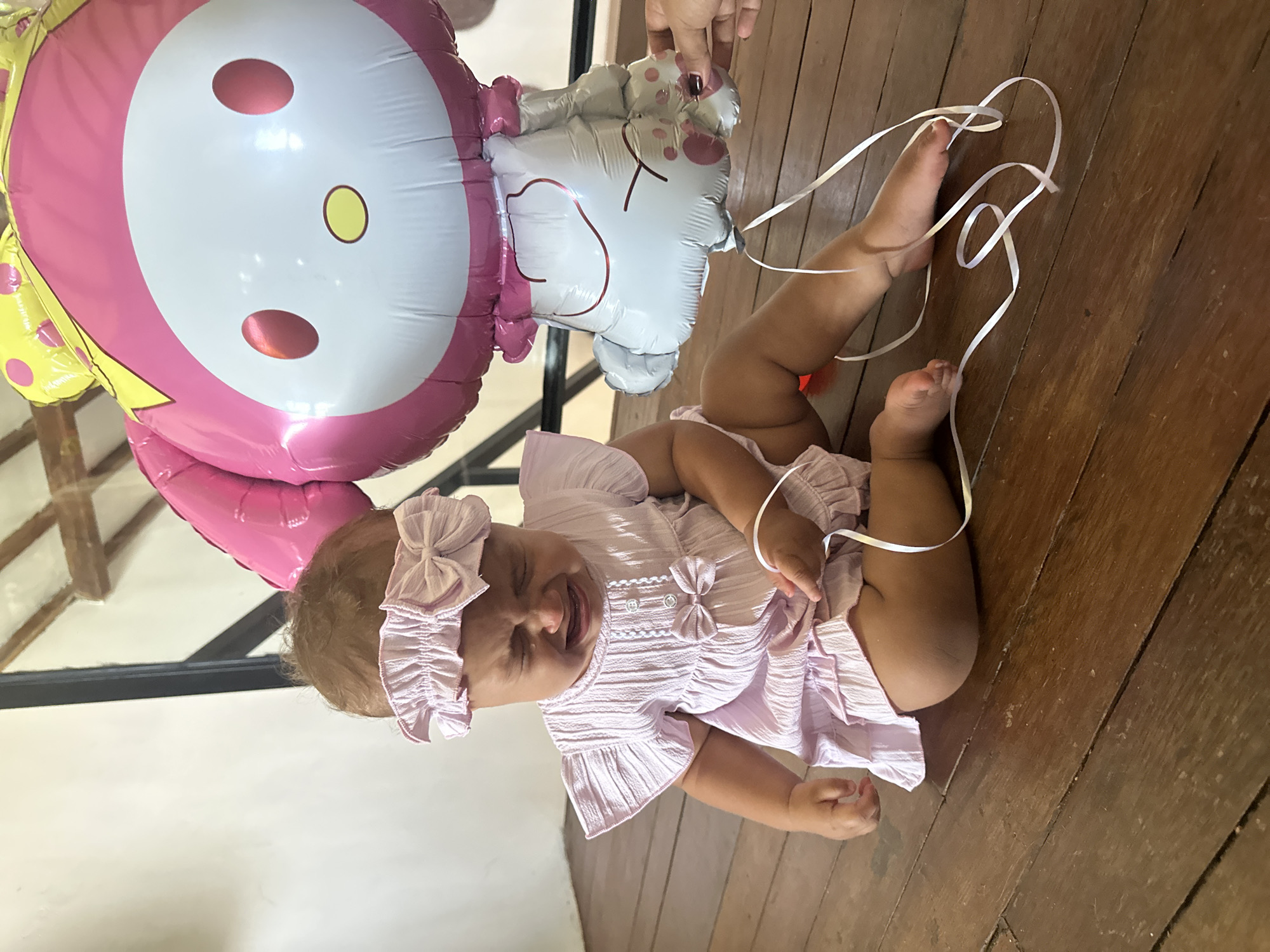 A crying baby is sitting on a wooden floor next to a My Melody balloon. The baby is wearing a pink dress and headband. A person's hand is holding the balloon string. The scene is indoors with wooden walls and a ceiling visible in the background. The location appears to be a residential area in North Kuta, Indonesia.