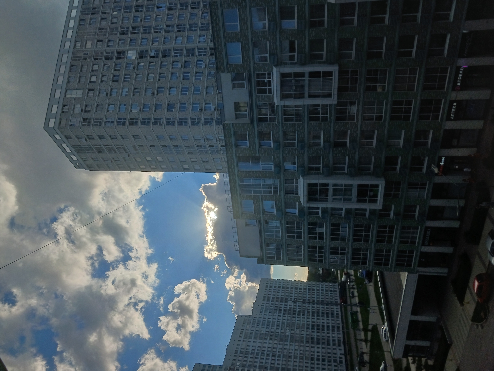 This is a photograph of a modern urban landscape in Yekaterinburg, Russia, taken from a low angle, looking upwards. The dominant subjects are two large, multi-story apartment buildings, one lighter in color and the other darker. The lighter building, on the left and in the foreground, has a uniform facade with numerous windows. The darker building, to the right, features a more textured facade with varied window sizes and protruding architectural elements.
The sky is a prominent element, with a mixture of bright blue sky and large, fluffy white clouds. Sunlight is breaking through the clouds, creating a lens flare effect and illuminating parts of the scene. A thin black wire or cable stretches diagonally across the sky. The overall weather appears to be partly cloudy and bright, suggesting a daytime setting, likely late morning or afternoon, given the intensity of the sunlight.
At the base of the darker building, there are visible signs of commercial activity. Text in Cyrillic script is legible on some of the storefronts, including "АПТЕКА" (APTEKA, meaning pharmacy) and what appears to be "ВкусВилл" (VkusVill, a Russian grocery store chain). Cars and possibly some people are visible on the street level, suggesting the buildings are in a residential or mixed-use area. The scene depicts everyday urban life, with no specific event taking place beyond the usual activities of a city. The composition emphasizes the scale of the architecture against the dynamic sky.