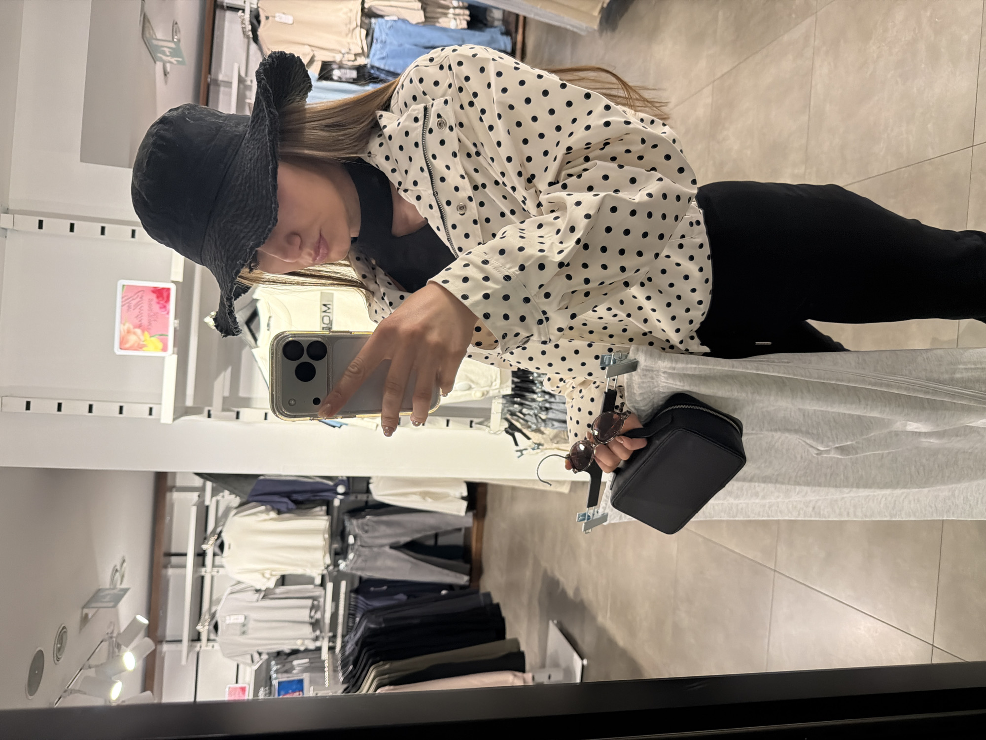 A woman is taking a selfie in a store, presumably in Sofia, Bulgaria, as that was specified in the prompt. She is wearing a black bucket hat, a white jacket with black polka dots, and black pants. She is holding a black purse and sunglasses. The reflection in the mirror shows racks of clothing. The store appears to be a clothing retailer.