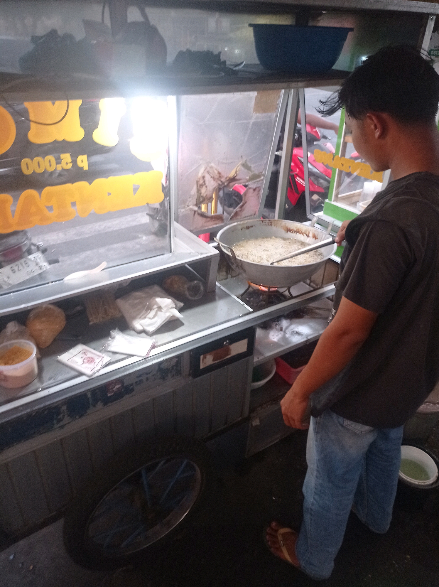 In Tuban, Indonesia, a person stands by a food cart. The person, wearing a dark t-shirt and blue jeans, is facing away from the camera and appears to be cooking. A large wok filled with oil and food is placed on a burner, and the person is holding a utensil over it. The food cart is stocked with various items, including packaged goods, containers of food, and what appears to be paper napkins. A blue basin is visible on a shelf above the cart. In the background, a motorcycle is parked behind the cart, and some buildings can be seen. The overall scene suggests a street food vendor operating in a bustling urban environment.