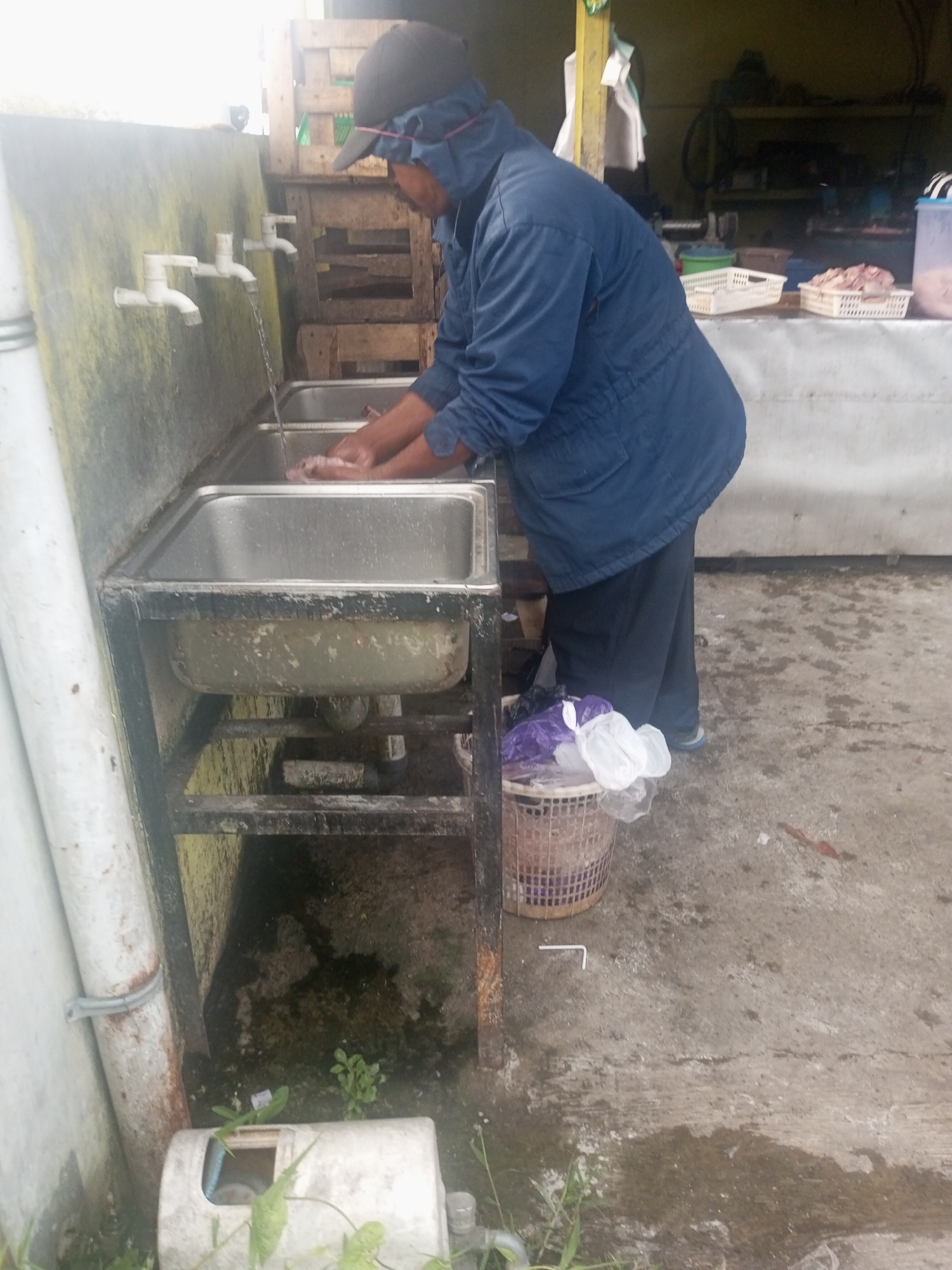 In Kalibeber, Indonesia, a person wearing a blue jacket and a black cap with a blue hood is washing their hands in a double-basin stainless steel sink. The sink is supported by a rusty metal frame and is located outdoors, next to a weathered, possibly moss-covered wall. Water flows from two white faucets mounted on the wall into the left basin. In the background, there are crates, shelves with white baskets containing food items, and a trash can overflowing with plastic bags. The ground is concrete and appears wet in places, with some debris and vegetation.